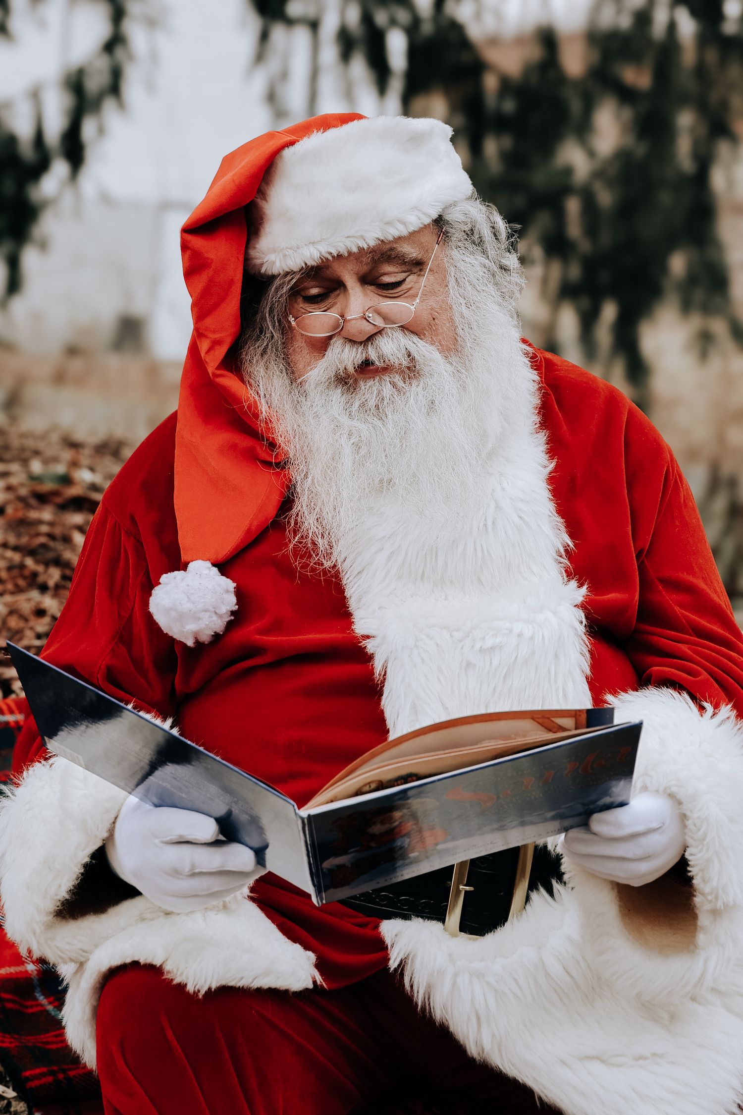 A person in a Santa Claus costume reads from a large book while sitting outdoors in a winter setting.