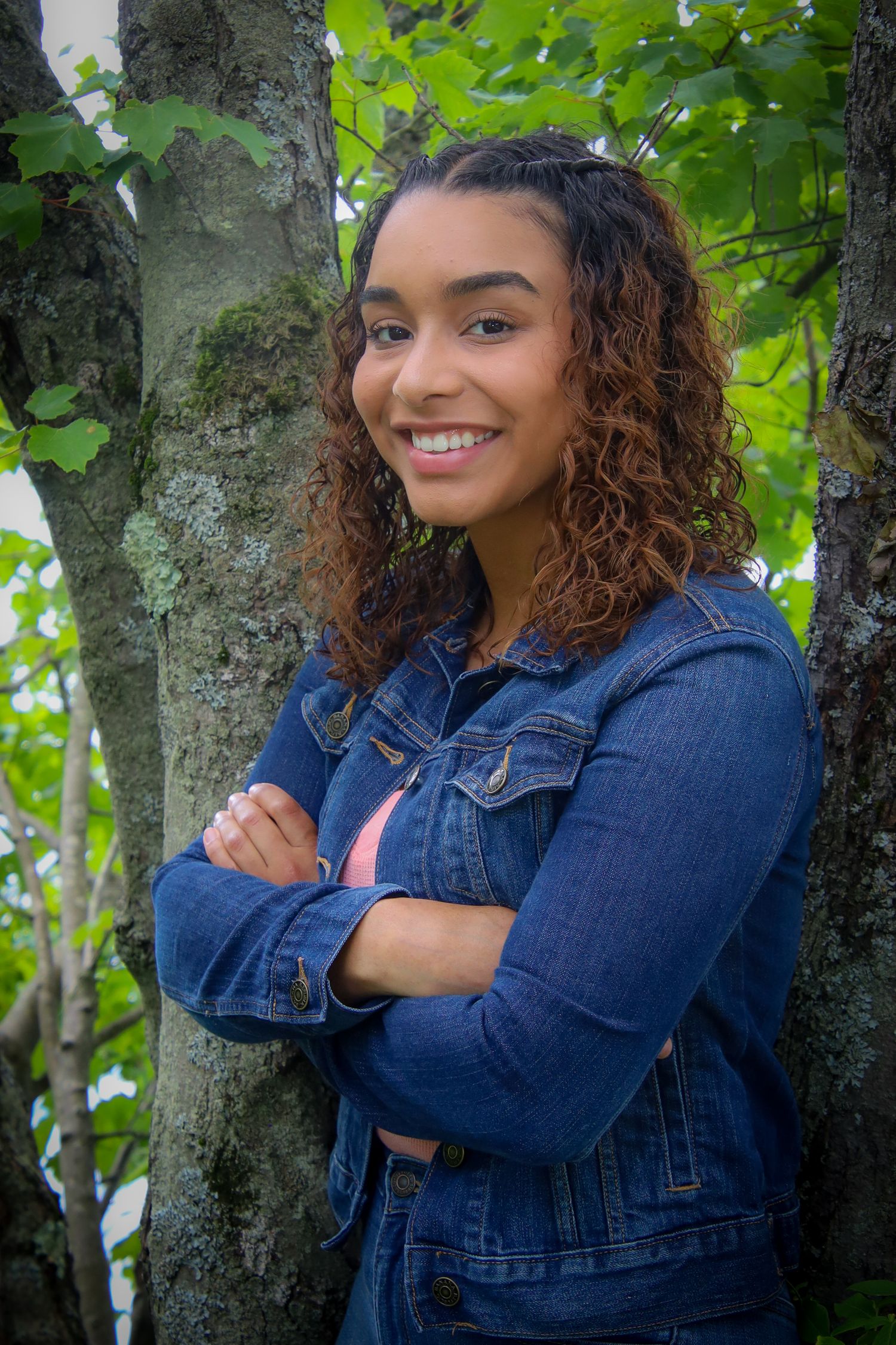 A person wearing a denim jacket stands with crossed arms by a tree trunk, with green leaves in the background.