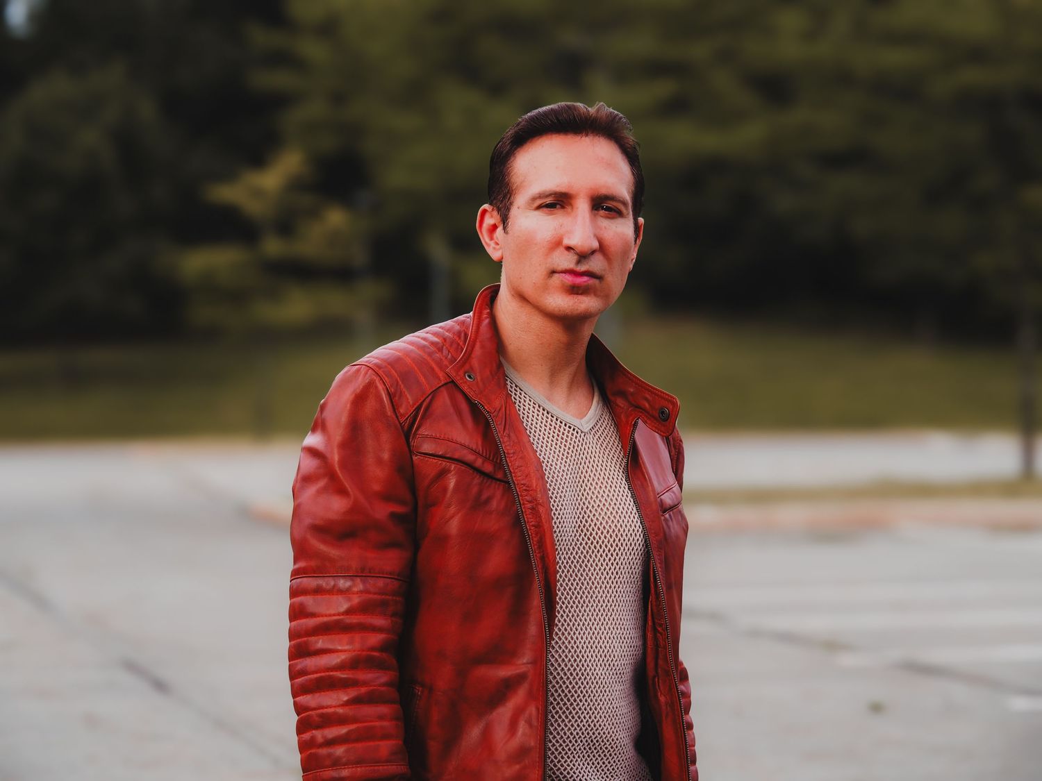 A person in a red leather jacket poses outdoors against a blurred natural background.