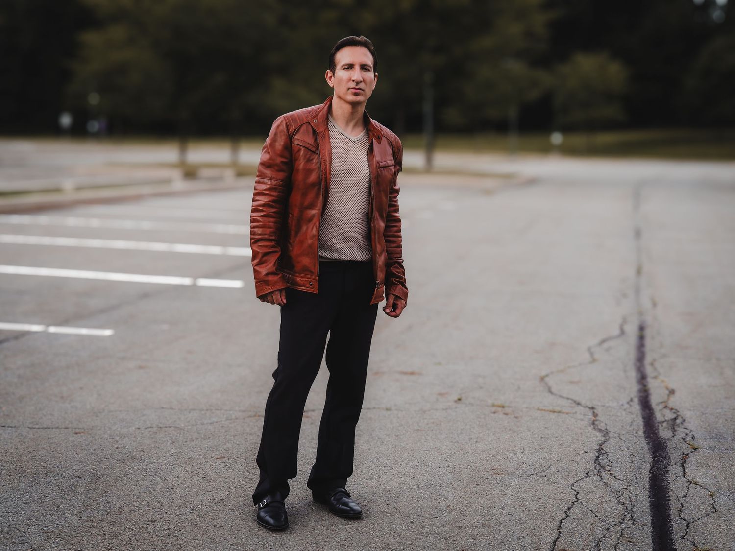 Person in brown leather jacket and dark pants standing in empty parking lot with tire marks on pavement.