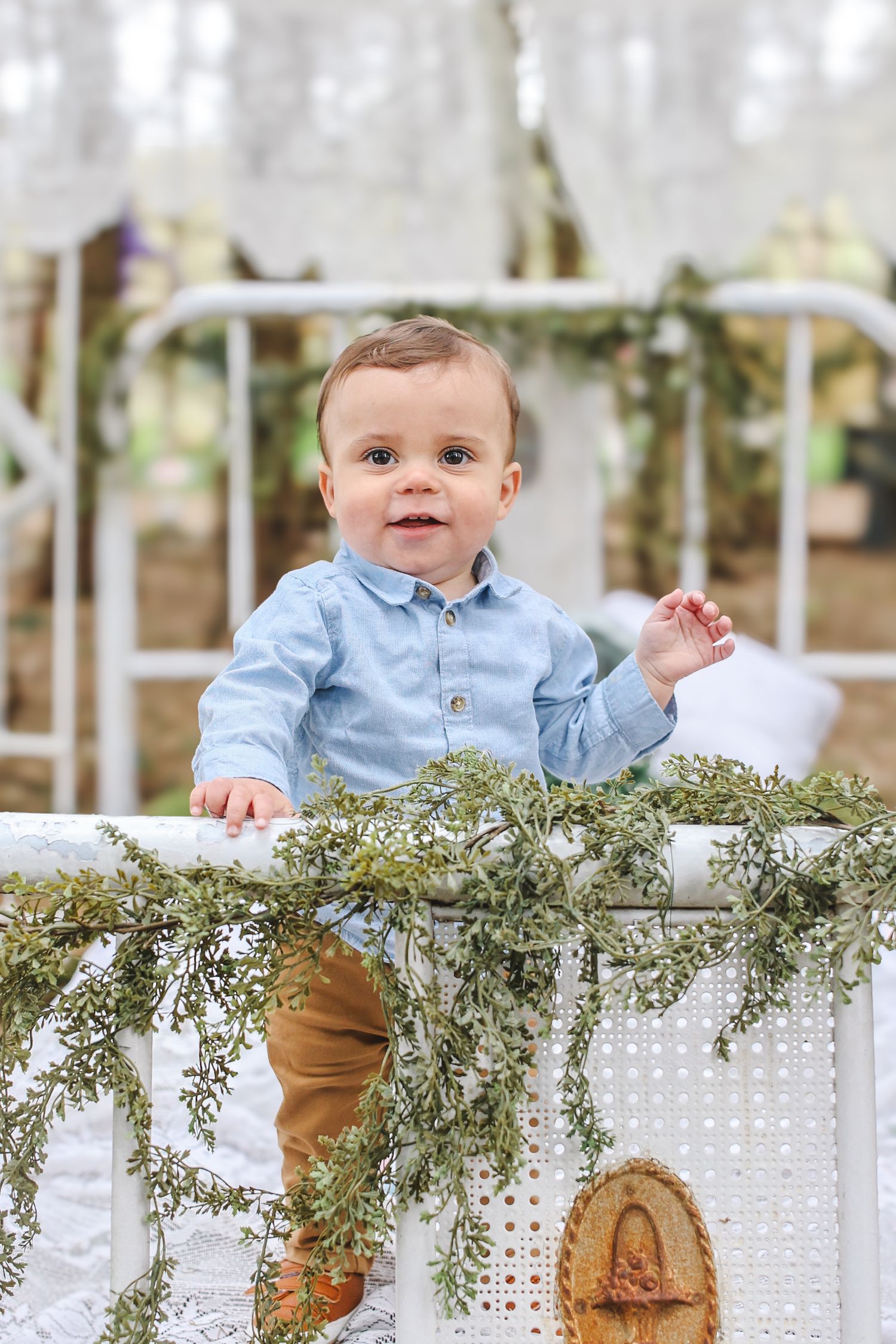 Baby in light blue shirt and brown pants smiles while standing at white fence decorated with greenery.