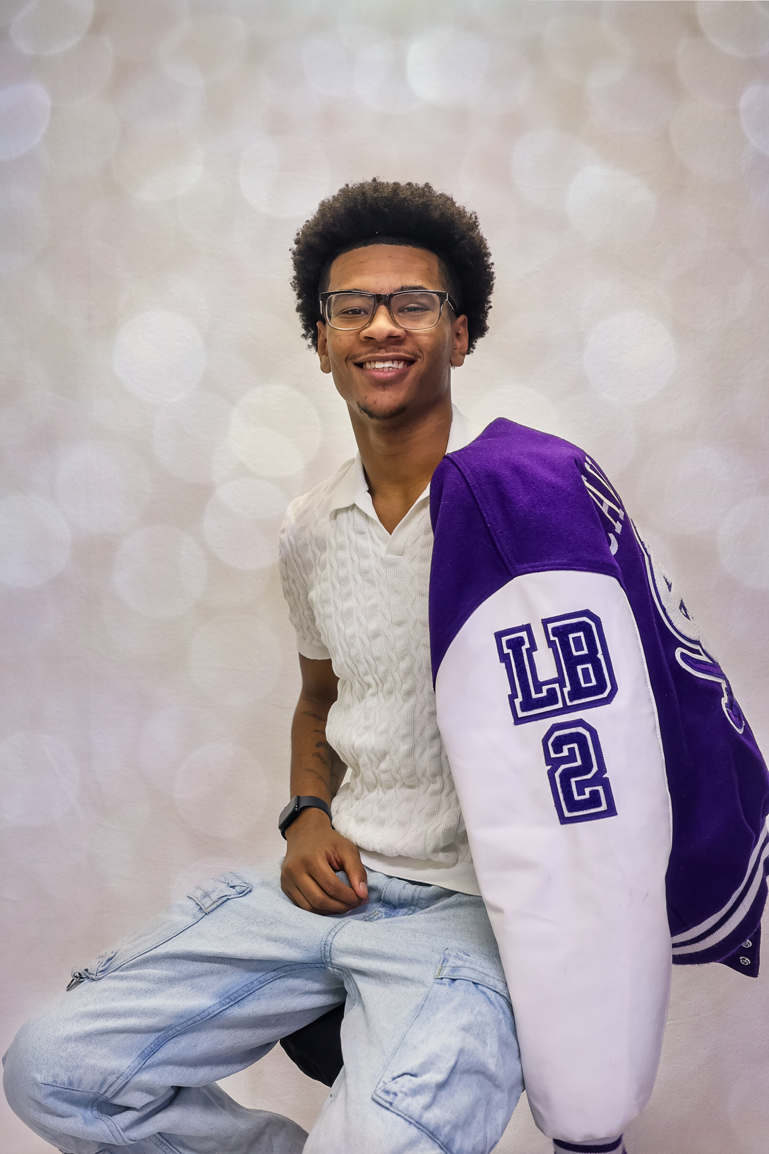Person in glasses wearing a purple and white varsity letterman jacket with light blue jeans in a studio setting.