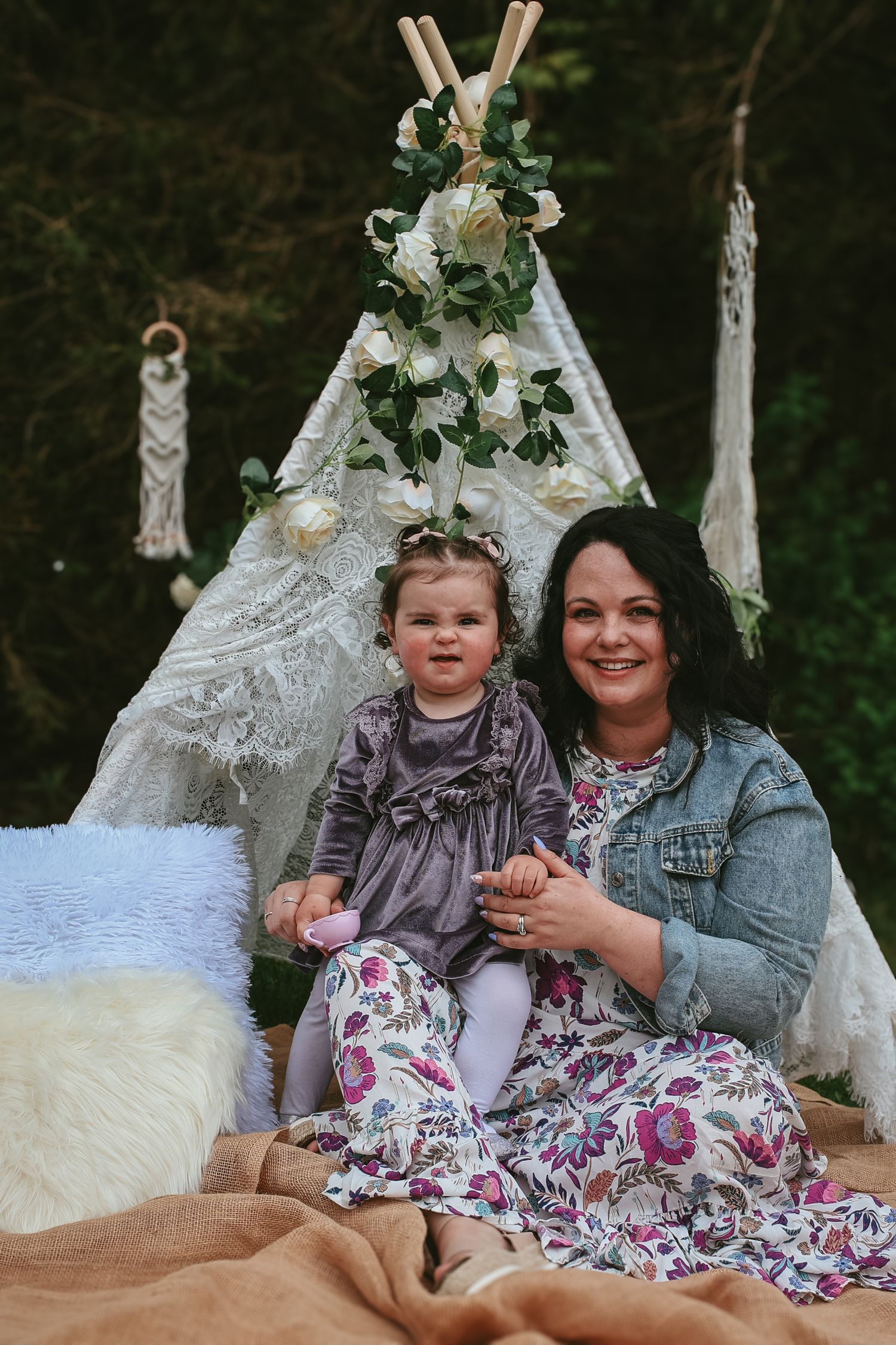 Decorative boho-style teepee setup with macrame hangings and floral arrangements in an outdoor woodland setting.