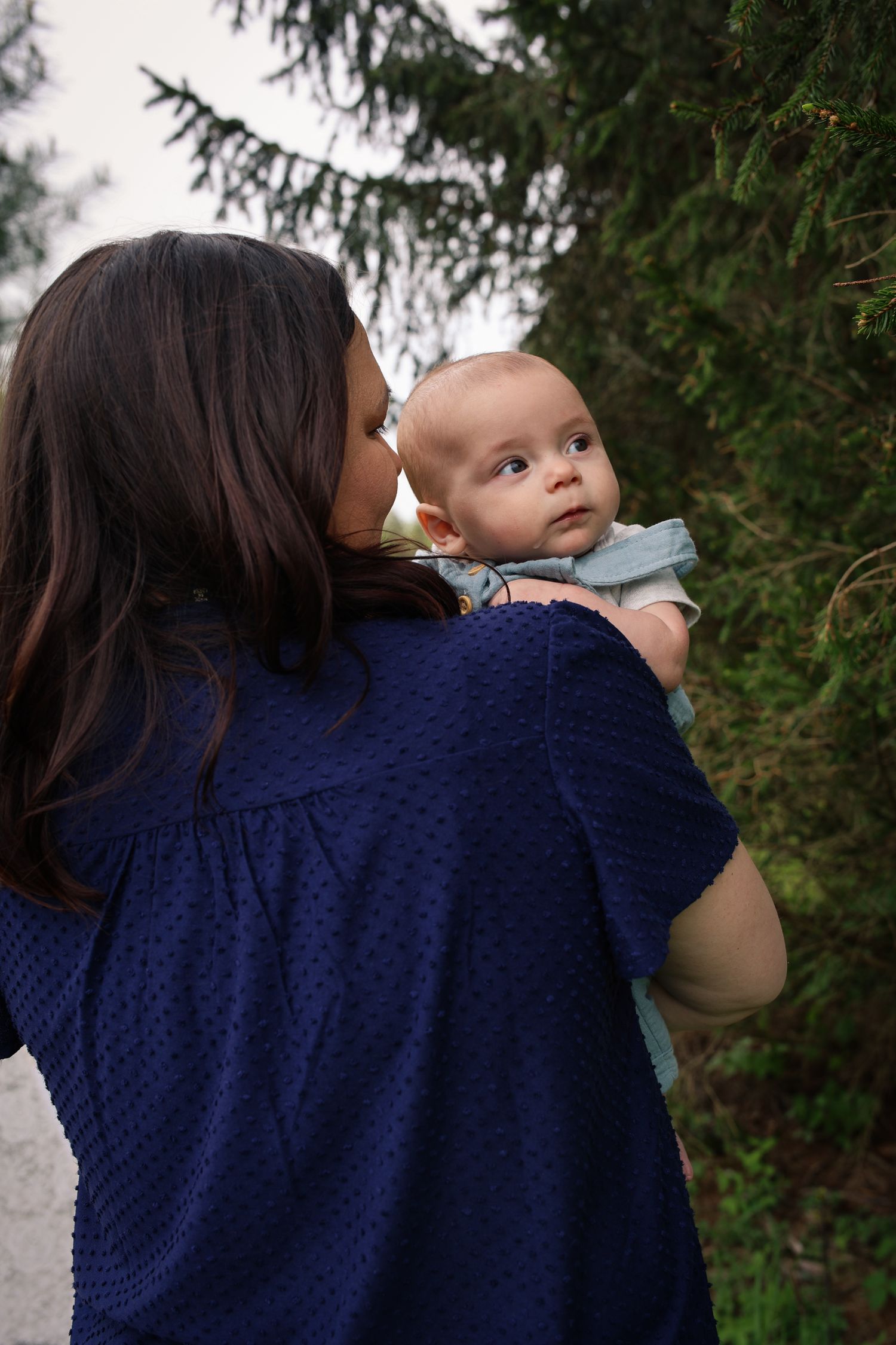 Parent in navy blue polka dot dress holds and comforts young baby in outdoor setting with evergreen trees.