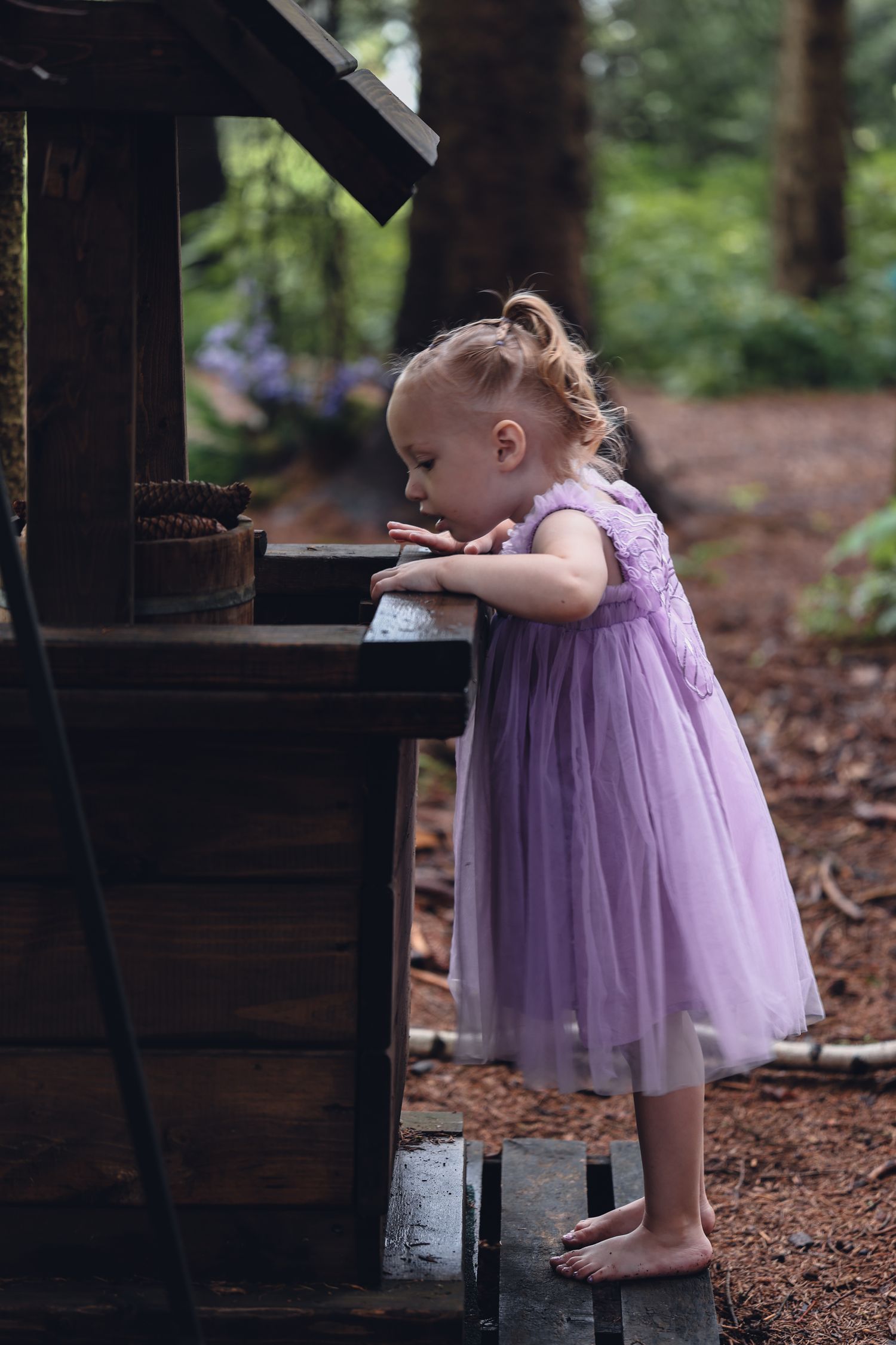 A small child in a purple tulle dress stands barefoot at a rustic wooden piano outdoors in a forest setting.