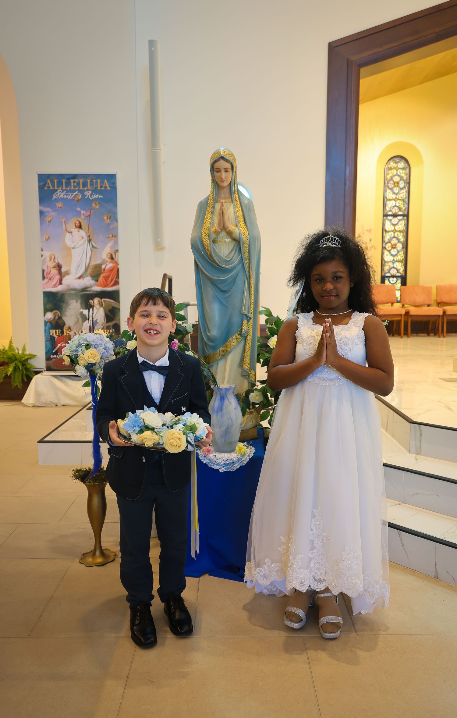 Two children in formal attire stand beside a religious statue of Mary in a church setting.