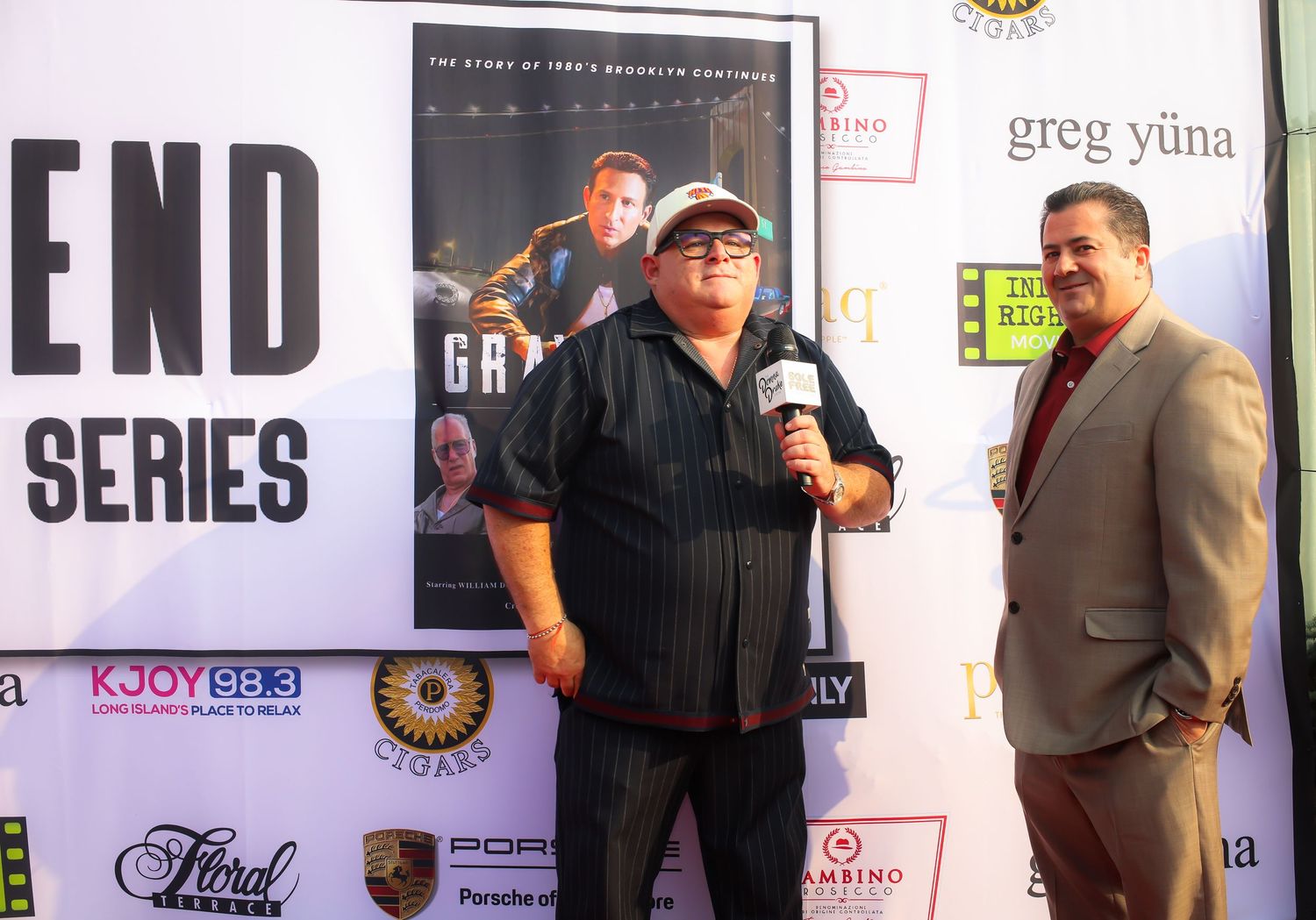 Three people stand together at a media event with promotional banners and signage visible in the background.