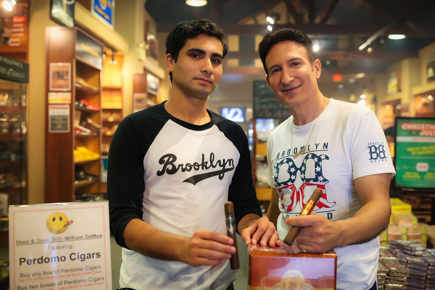 Two people smile while working behind the counter at a cigar shop with shelves of products visible in the background.