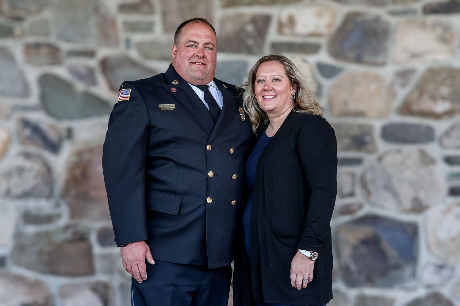 A couple poses together in front of a stone wall, with one wearing a formal dress uniform and the other in black attire.