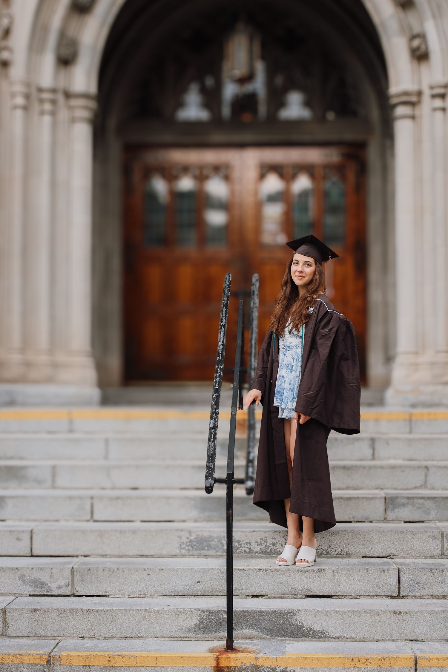 A graduate in cap and gown poses for portraits on stone steps of a grand academic building.