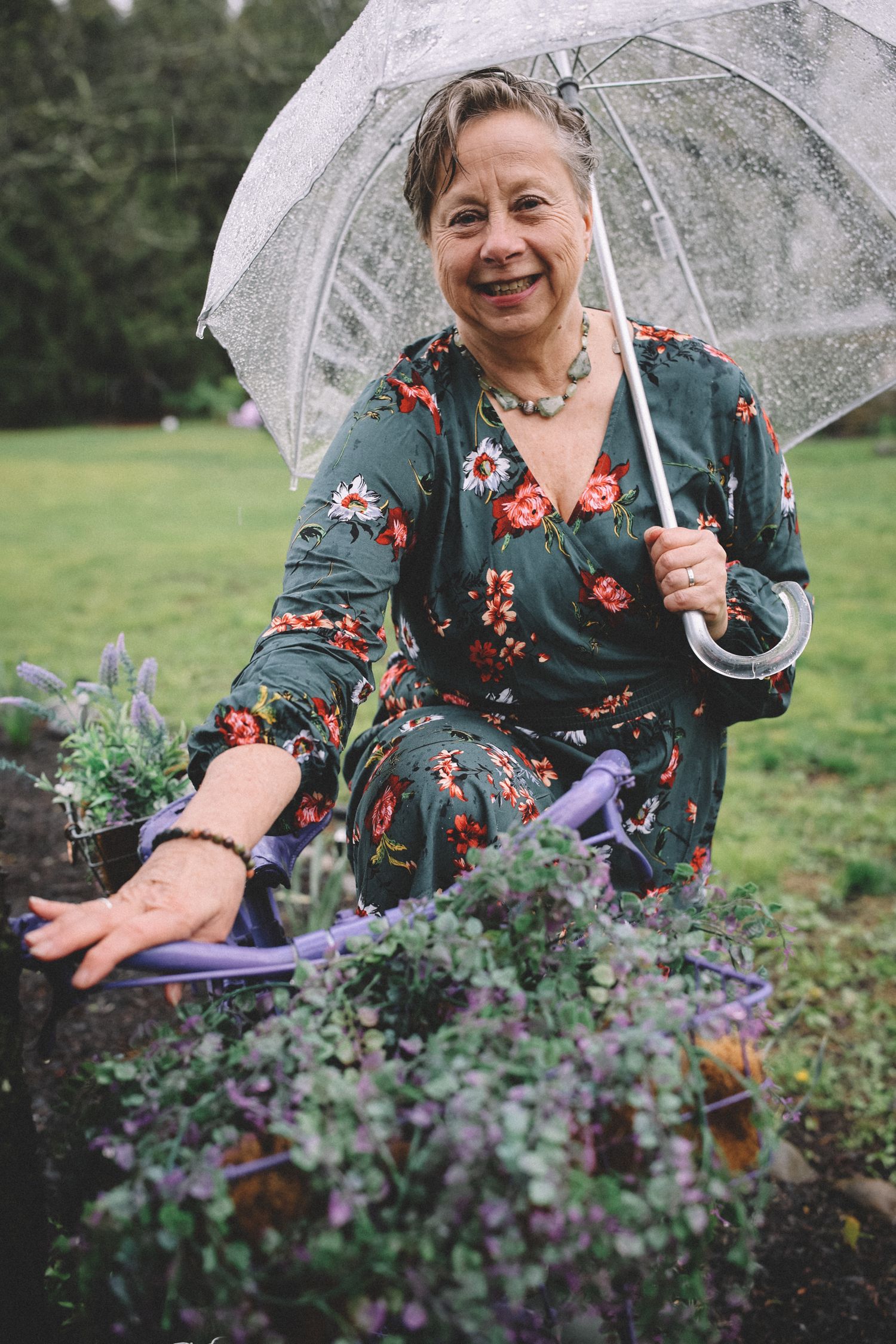 Someone in a floral dress holds a clear umbrella while sitting in a garden with purple flowers on a rainy day.