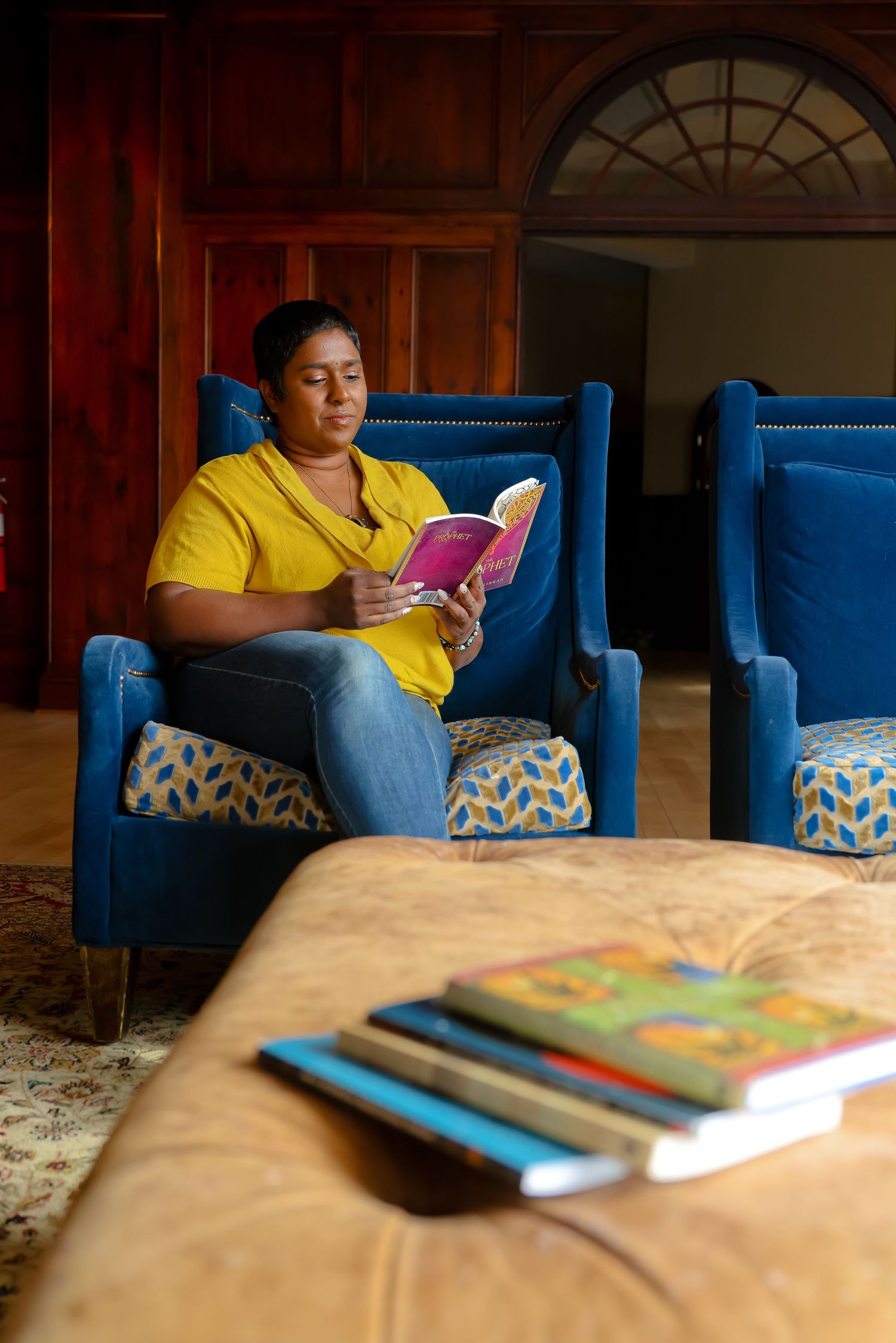 Person in yellow shirt sits on blue chair reading book in warm-lit room with books on wooden table.