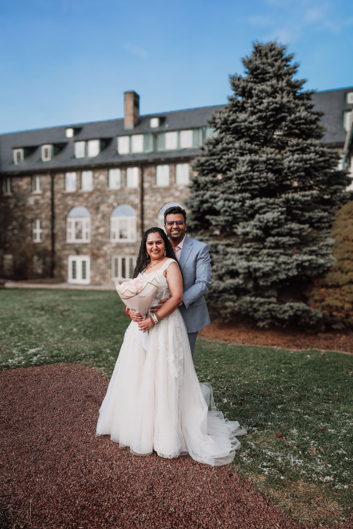 A couple poses for a wedding photo in front of a stone building with evergreen trees on a sunny day.