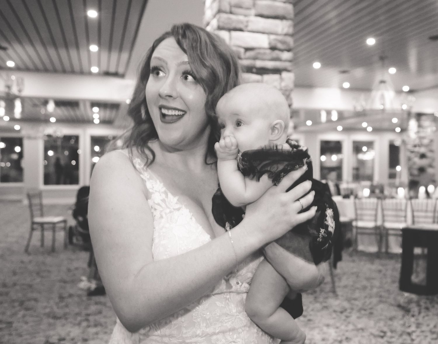 A bride in a white dress holds and dances with a small black puppy at an indoor wedding reception.