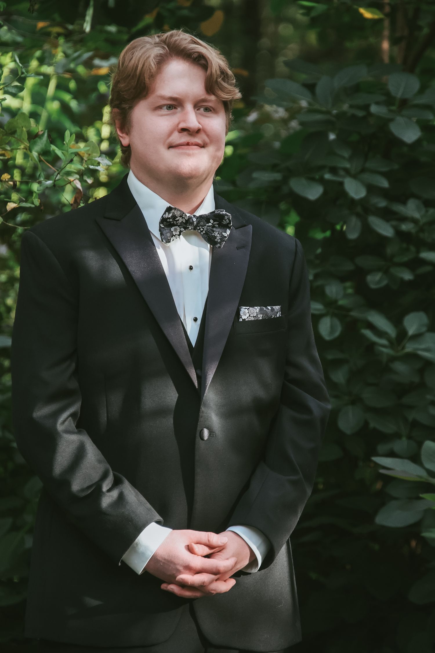 A person in formal black tuxedo attire stands in front of lush green foliage during an outdoor photo session.