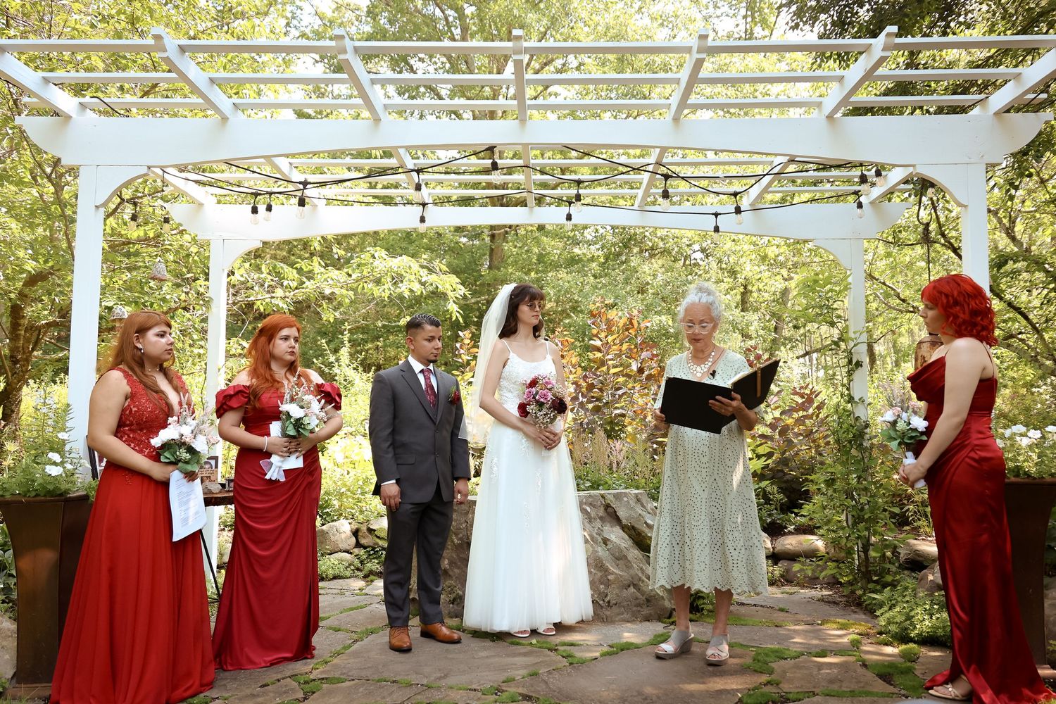 Wedding party members in red dresses and formal attire gather under a white pergola for outdoor celebration photos.