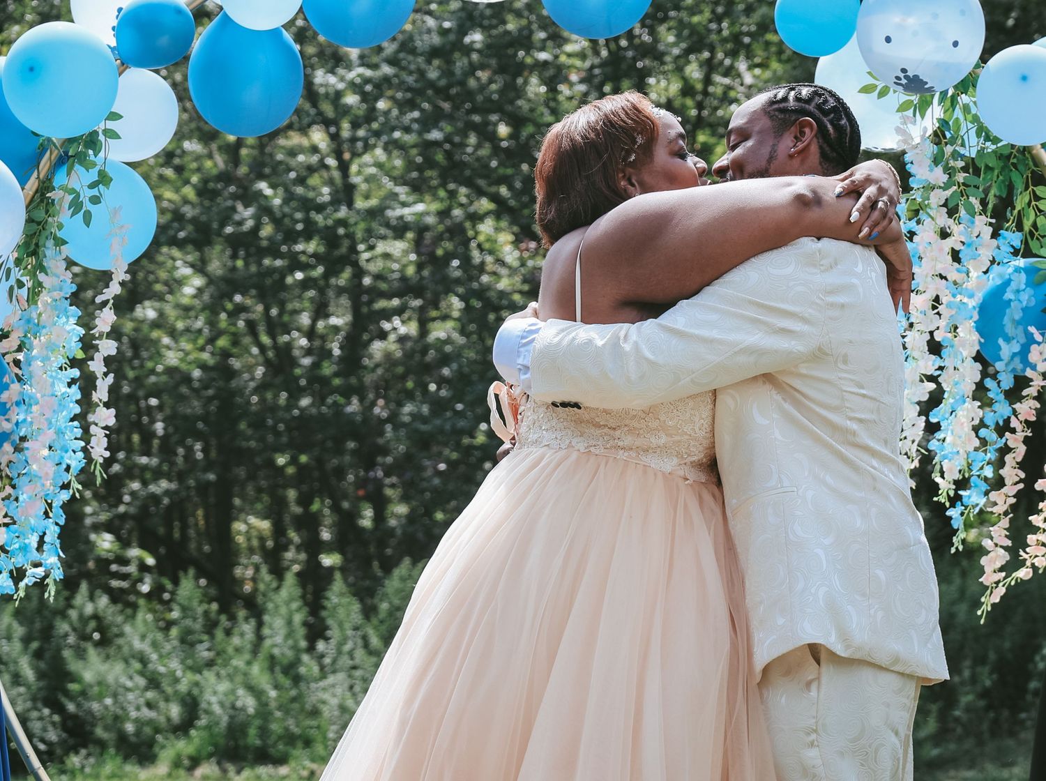 A romantic outdoor wedding scene with blue balloons floating in the air as a couple embraces under green trees.