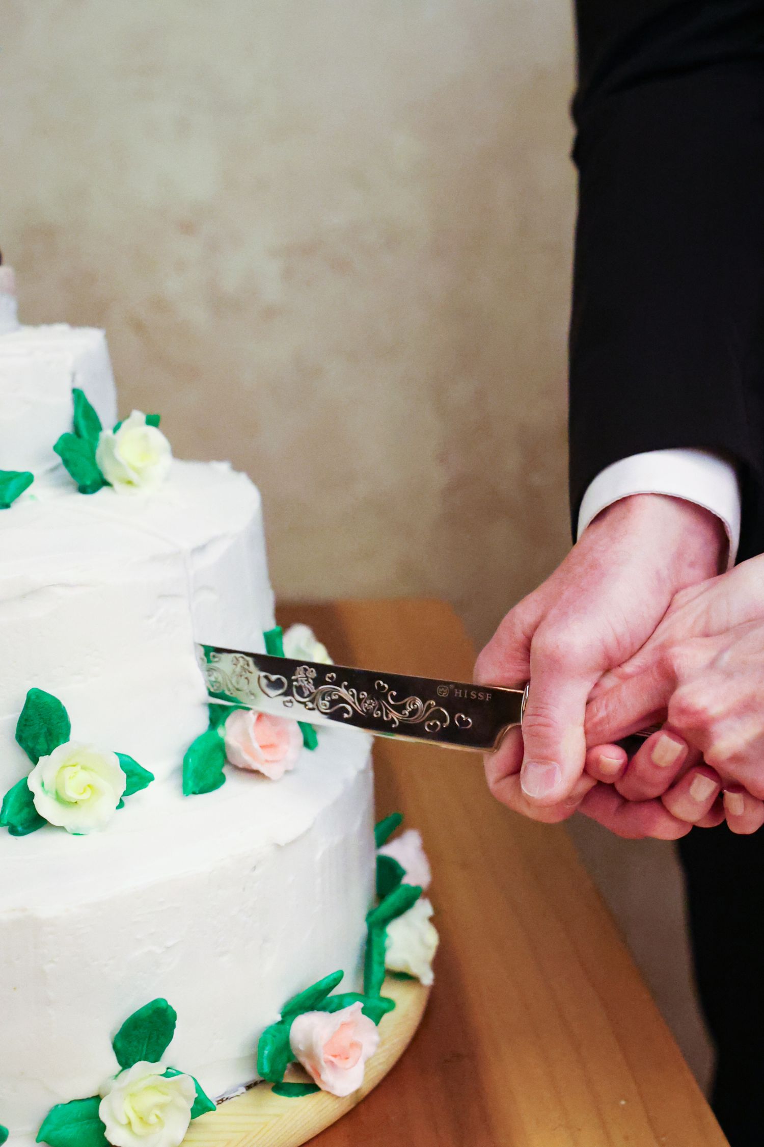 A white wedding cake with green leaves and pale roses is being cut with a silver knife on a wooden table.