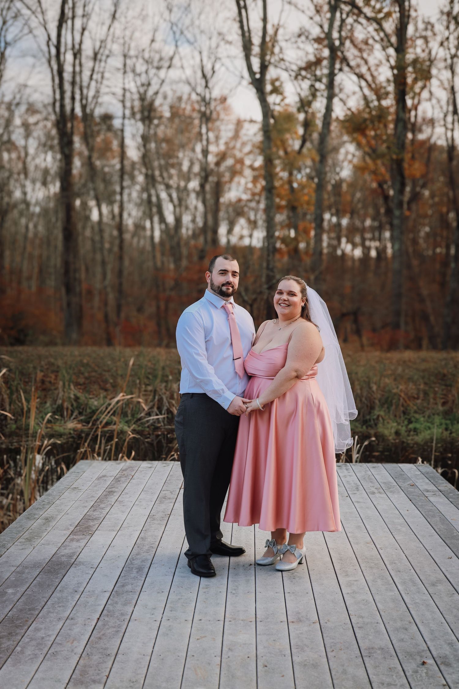 A couple stands on a wooden deck surrounded by autumn trees, wearing pink and blue formal attire for their wedding day.