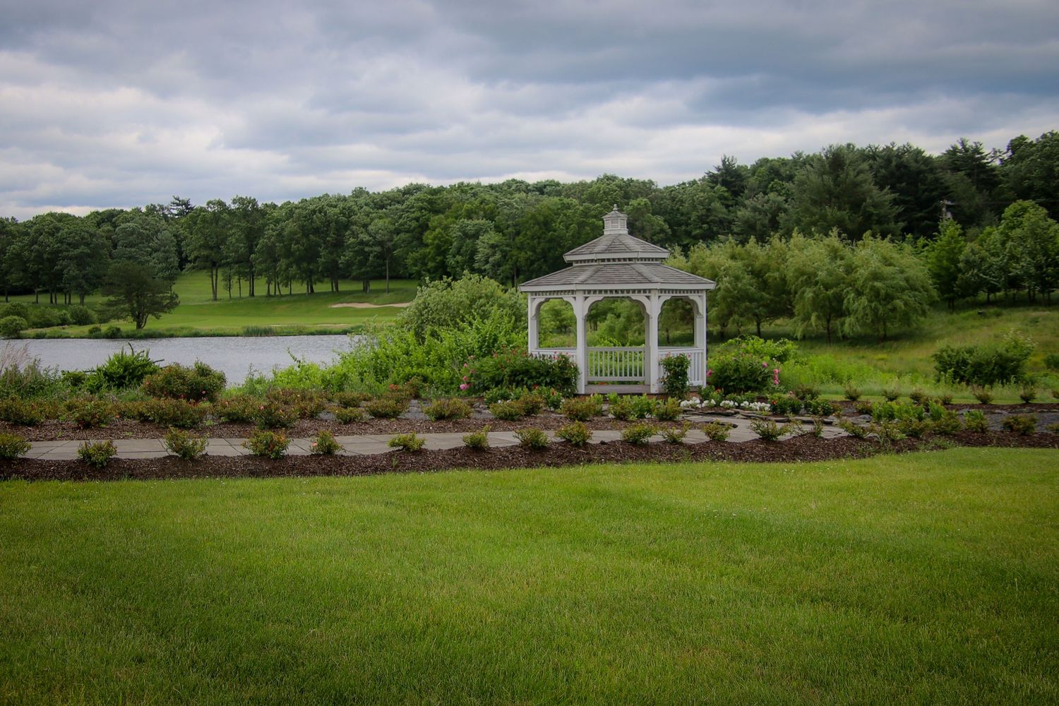 A scenic panoramic view of a white gazebo and landscaped gardens overlooking a tranquil lake surrounded by lush greenery.