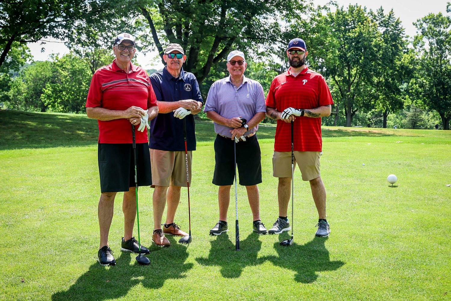 Four men in red polo shirts pose together on a sunny golf course holding clubs.