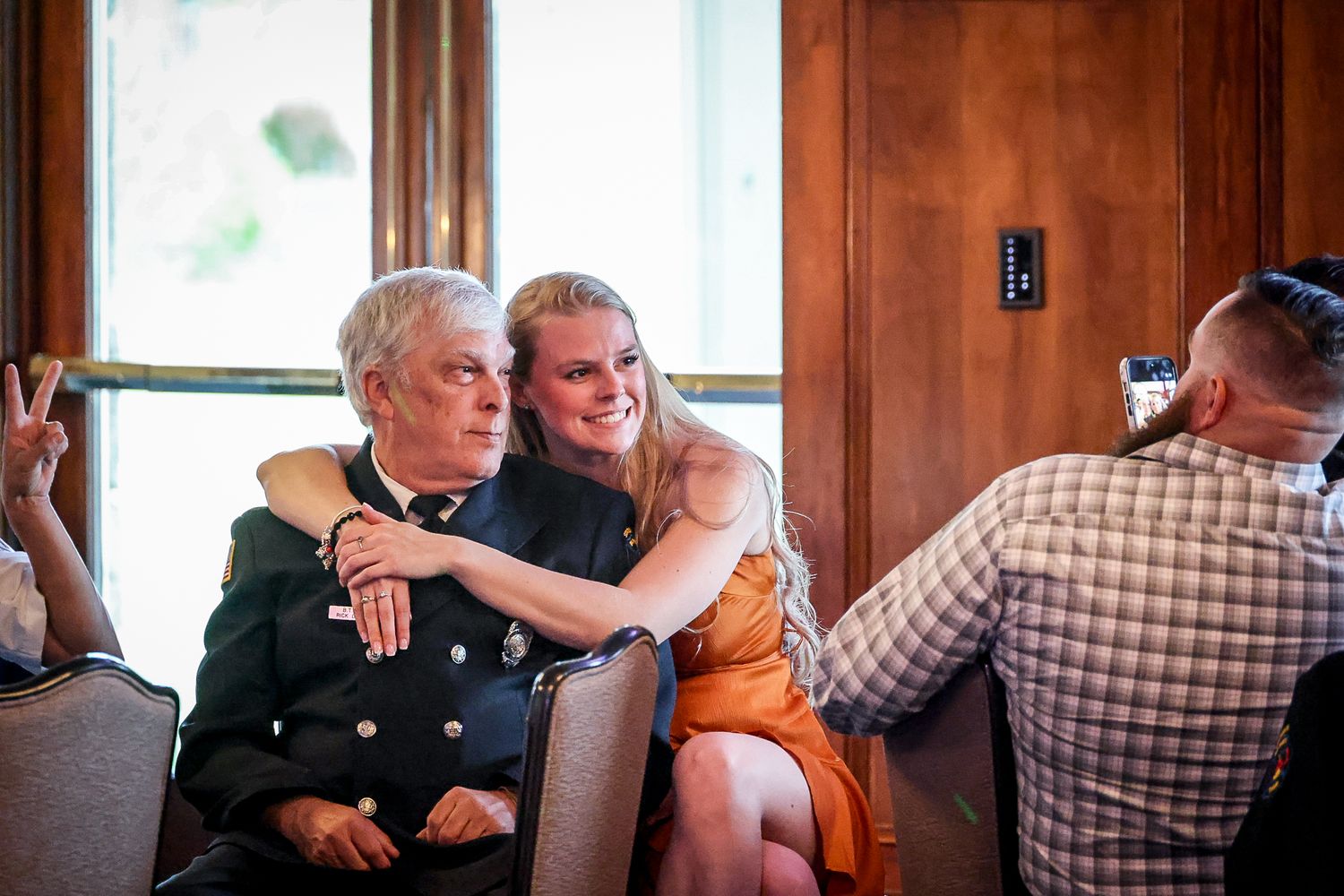 A young woman in an orange dress embraces an elderly person in a dark jacket at an indoor social gathering.