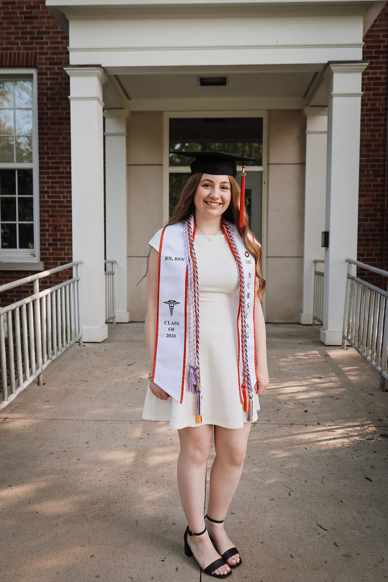 Smiling female graduate in cap and gown stole poses outdoors in front of brick building columns on graduation day.