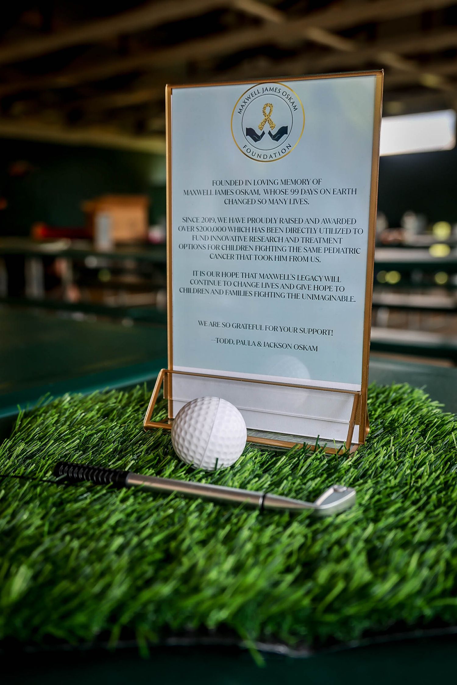 Gold-framed charity sign beside a golf ball and club on artificial green turf at a fundraising golf event.