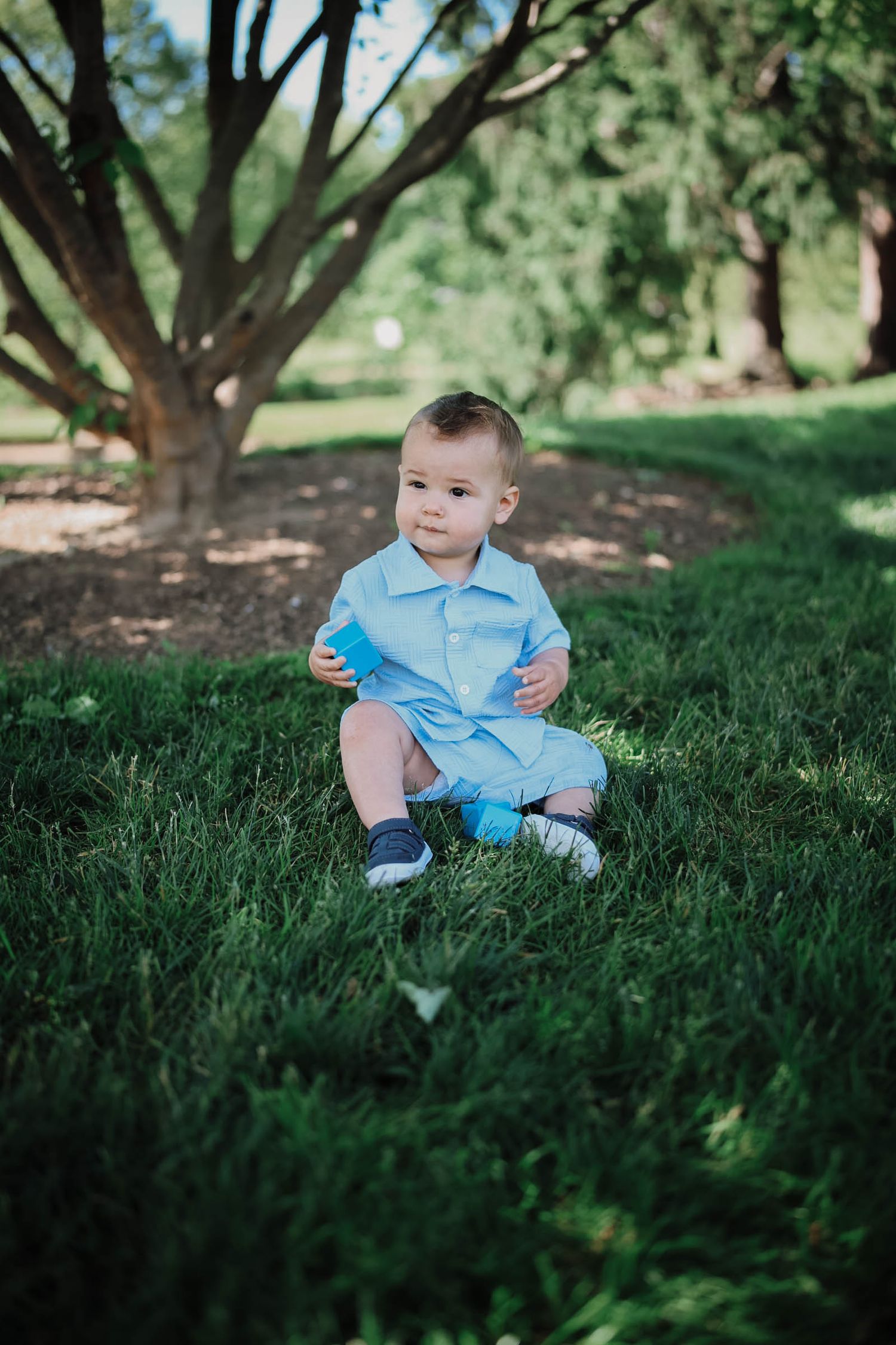 Adorable baby boy in blue romper sitting on lush green grass in a sunny park surrounded by trees.
