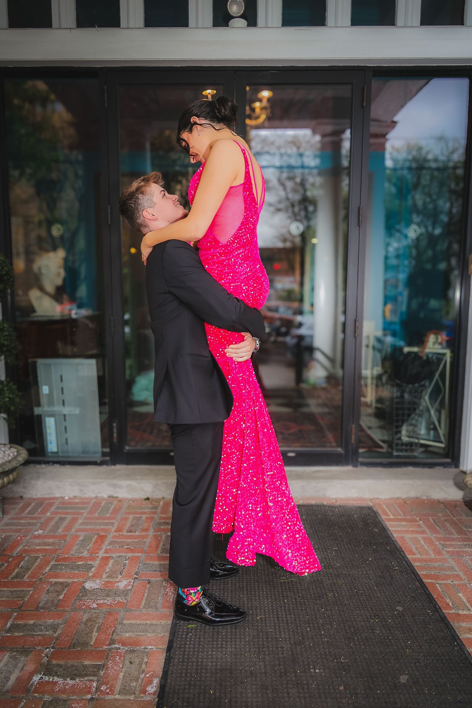 A man in a black suit lifts a woman in a sparkling pink gown outside a building entrance.
