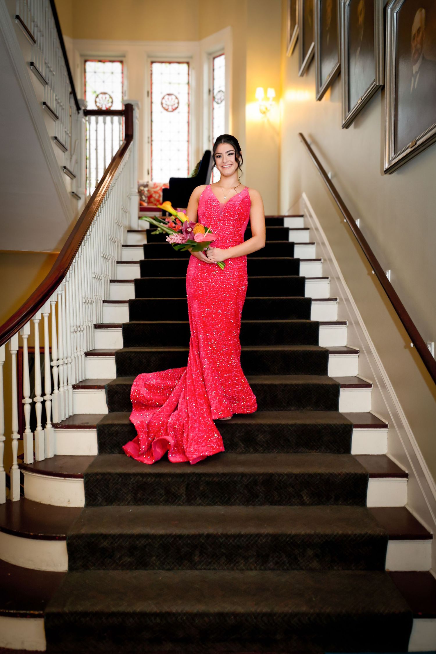 Woman in red sequin mermaid gown holding tropical bouquet poses elegantly on grand staircase.