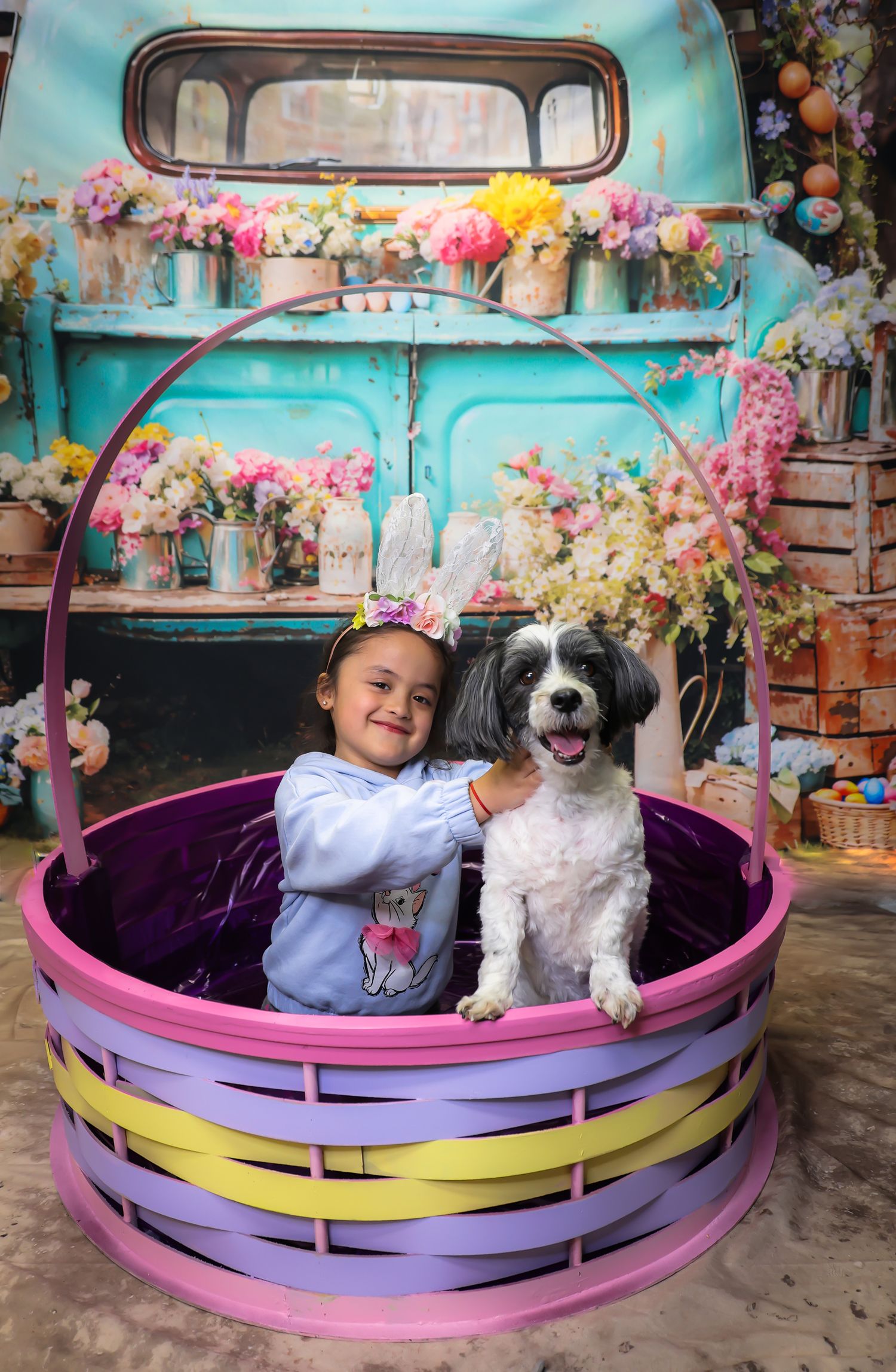 A young girl and a black-and-white dog sit together in a large Easter basket before a floral-decorated vintage teal truck.