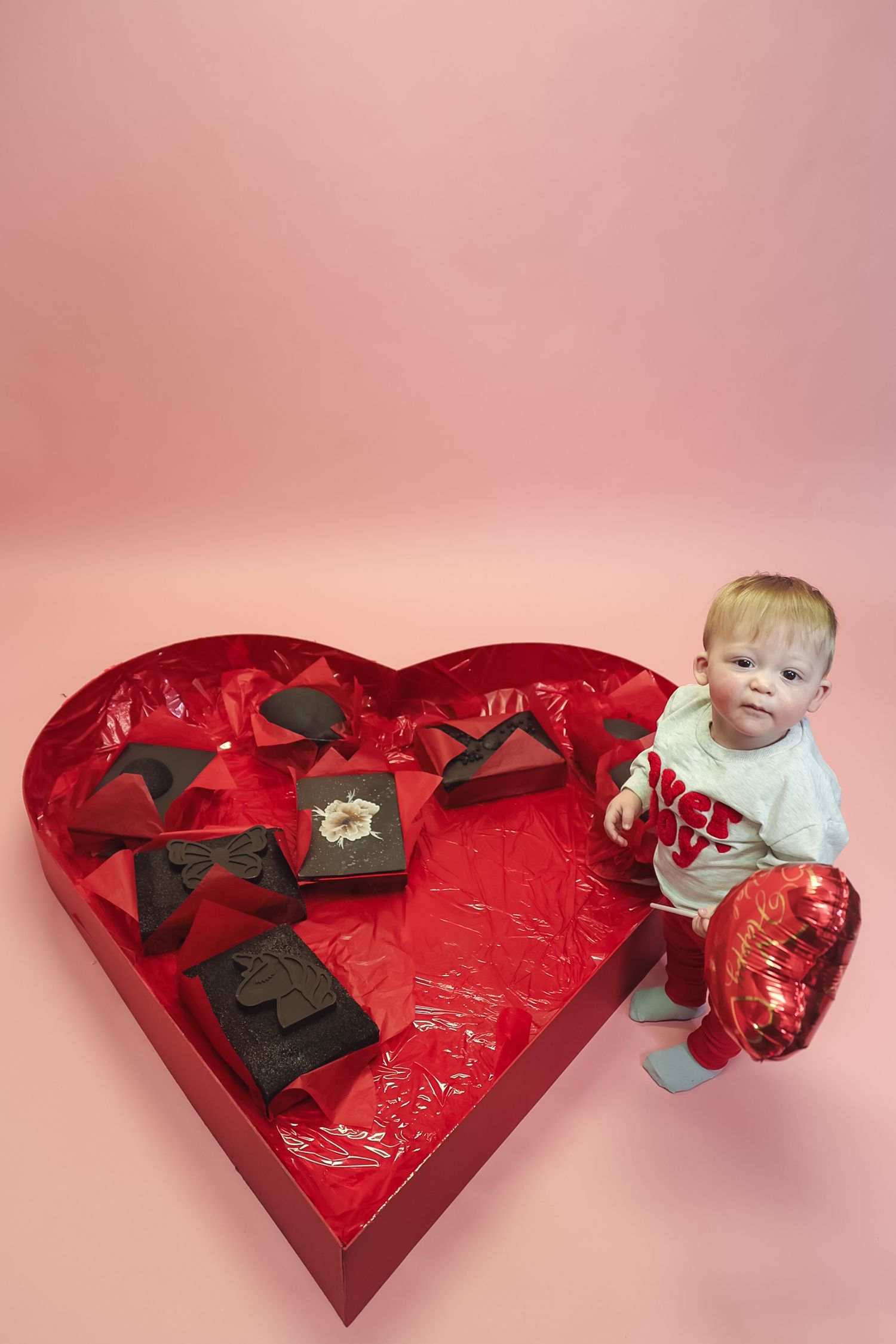 Toddler sitting in a large red heart-shaped Valentine's Day chocolate box on a pink background.