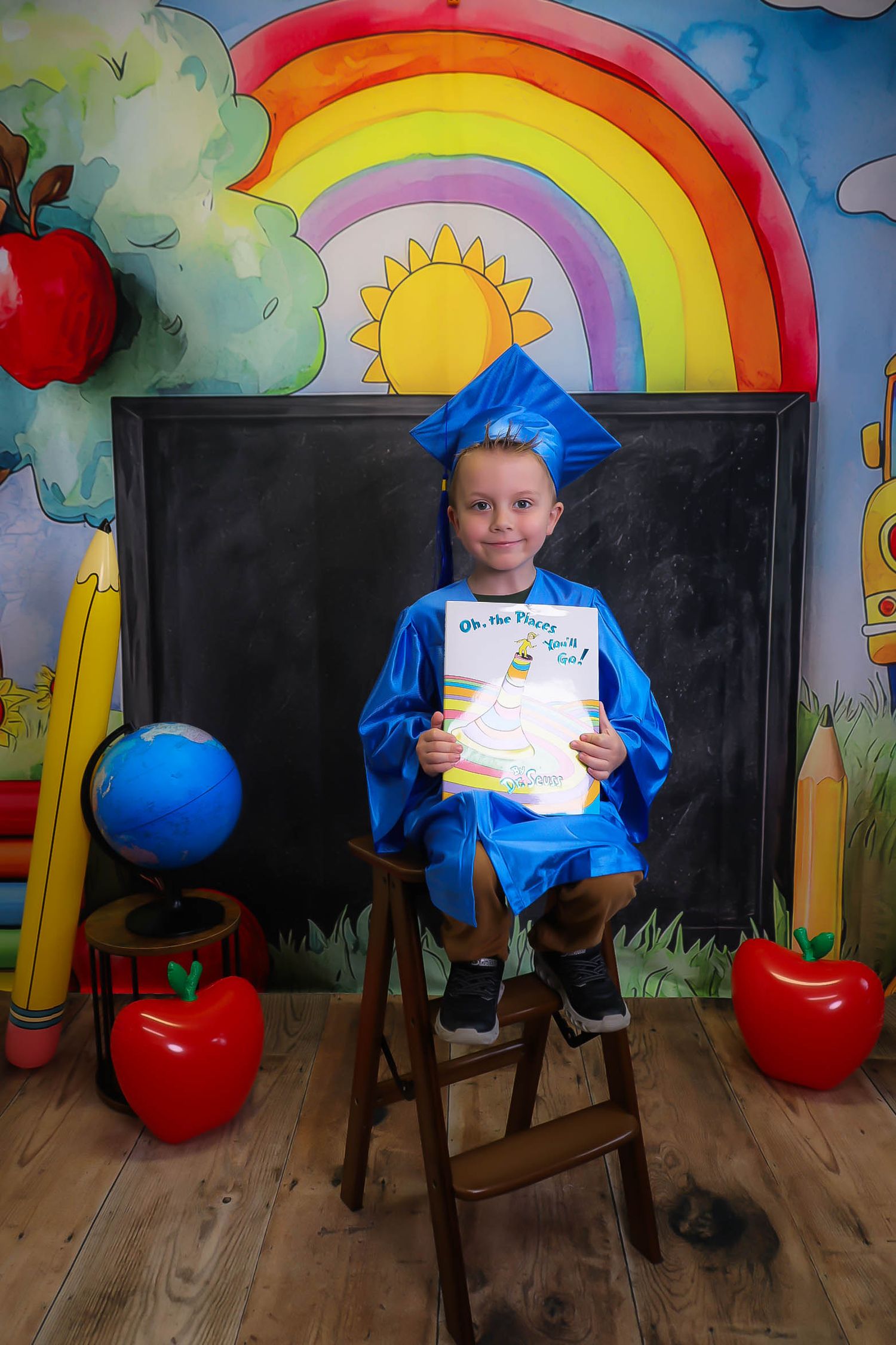 Young boy in blue graduation cap and gown sits on stool holding diploma in colorful classroom setting.