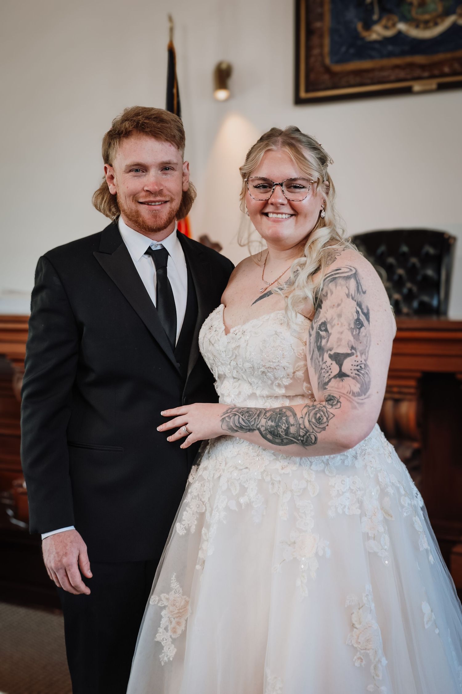 Bride in lace gown with tattoos and groom in black suit smiling together at a courthouse wedding ceremony.