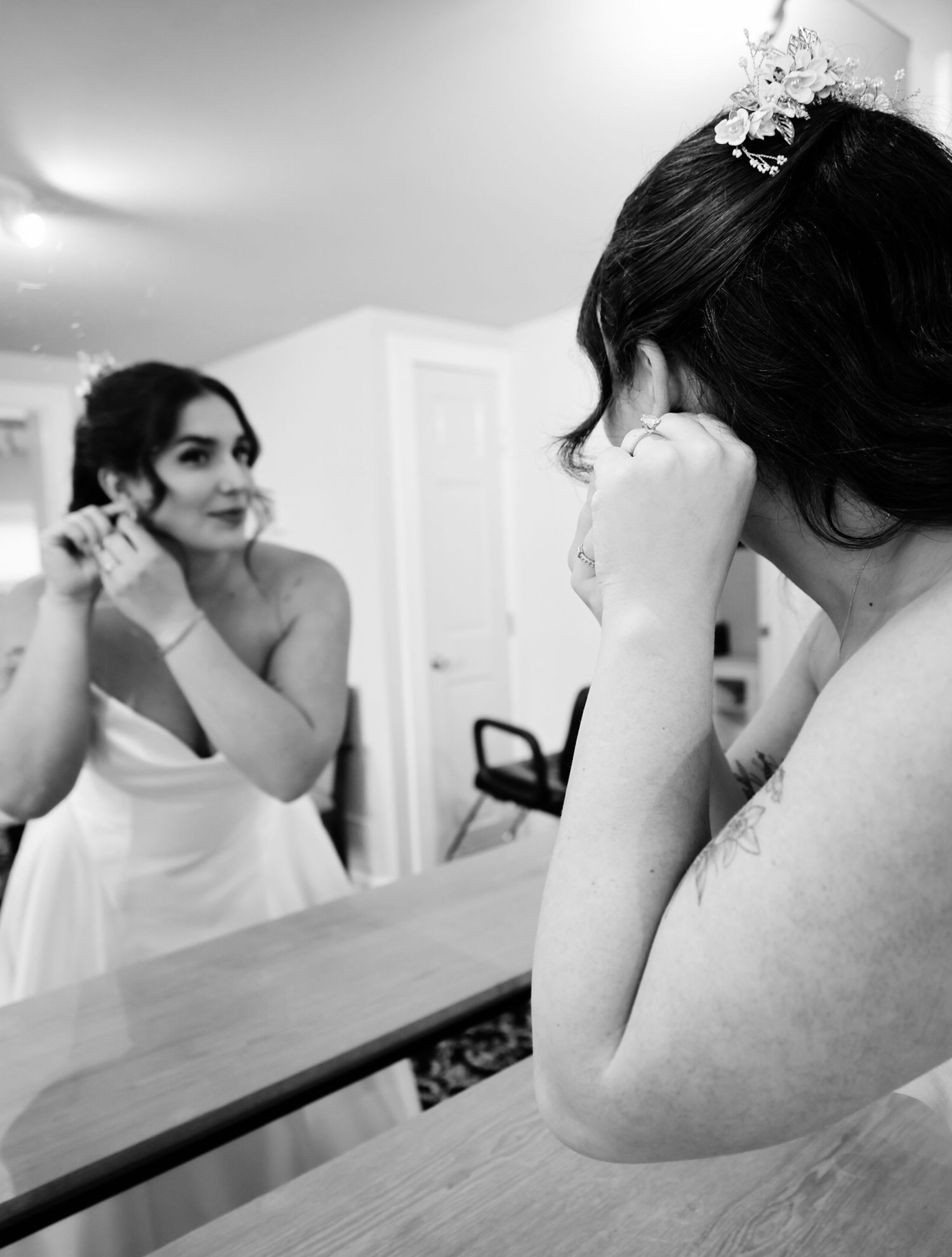 Bride in white gown adjusting earring while looking in mirror, black and white wedding photo.