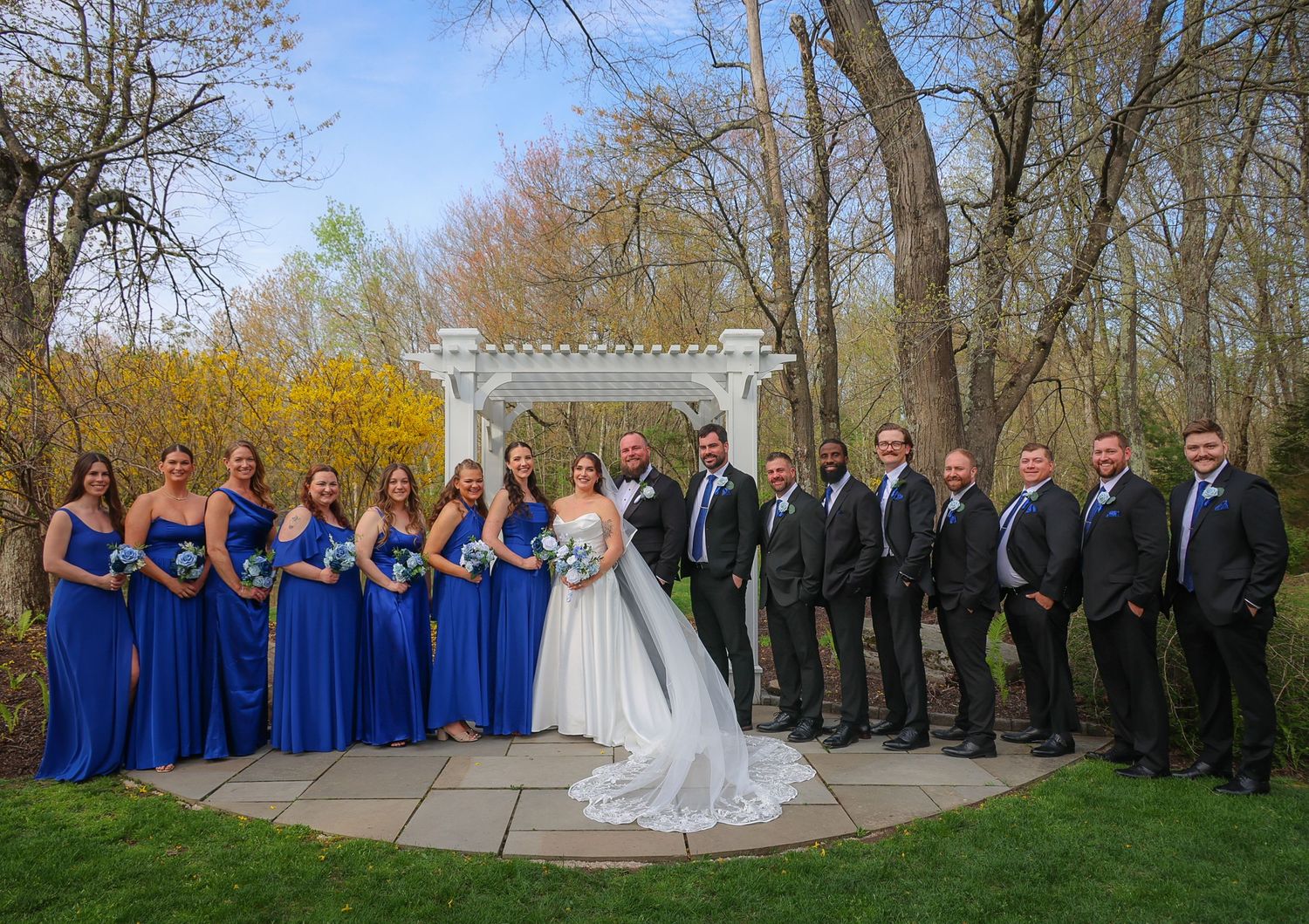 Wedding party poses outdoors near white gazebo, bride in white gown with bridesmaids in royal blue and groomsmen in gray suits.