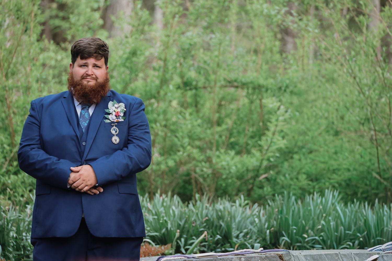 Bearded man in navy blue suit with boutonniere stands outdoors among lush green trees and plants.