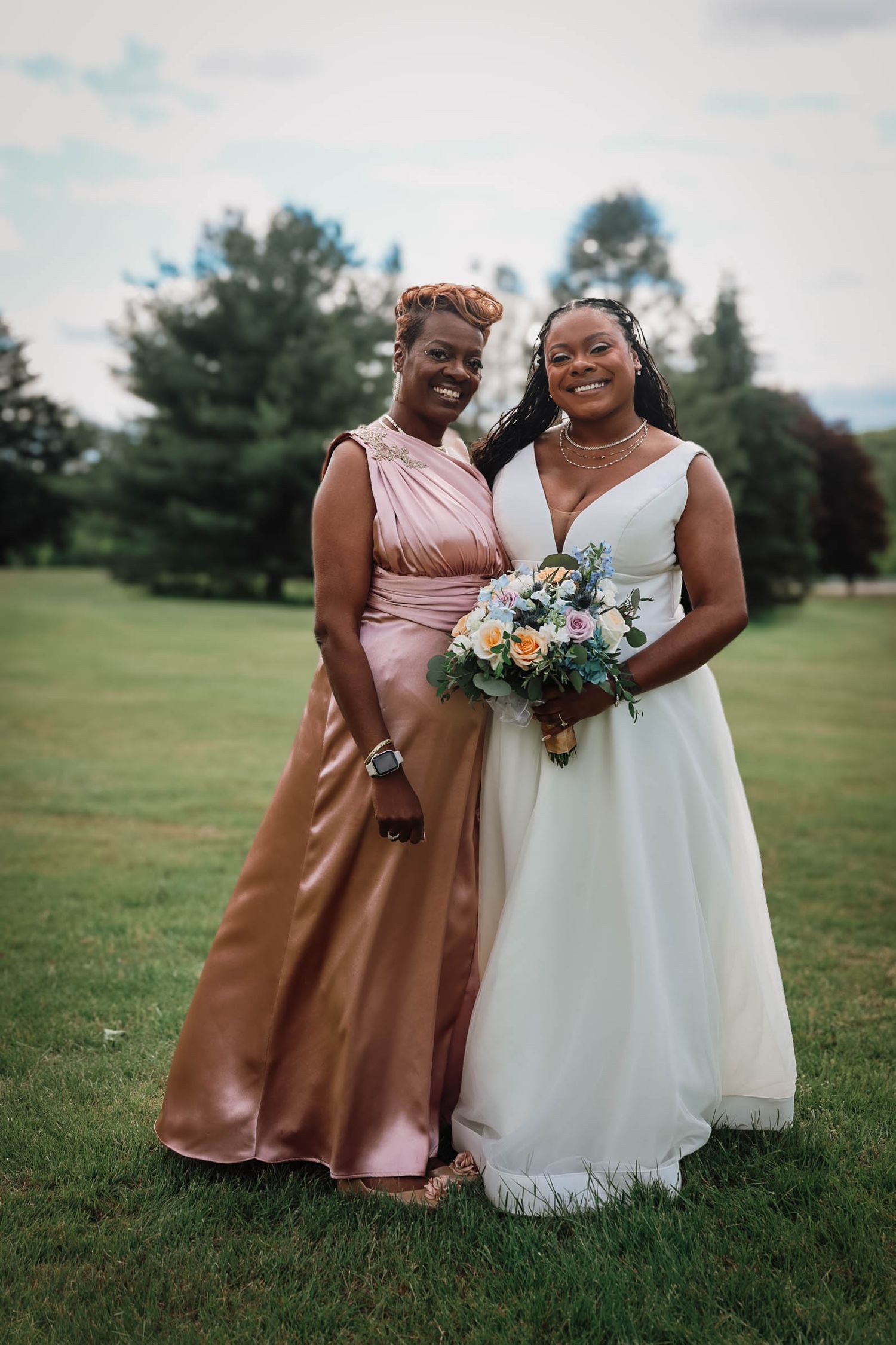 Bride in white gown holding bouquet poses with bridesmaid in rose gold dress on green lawn at outdoor wedding.