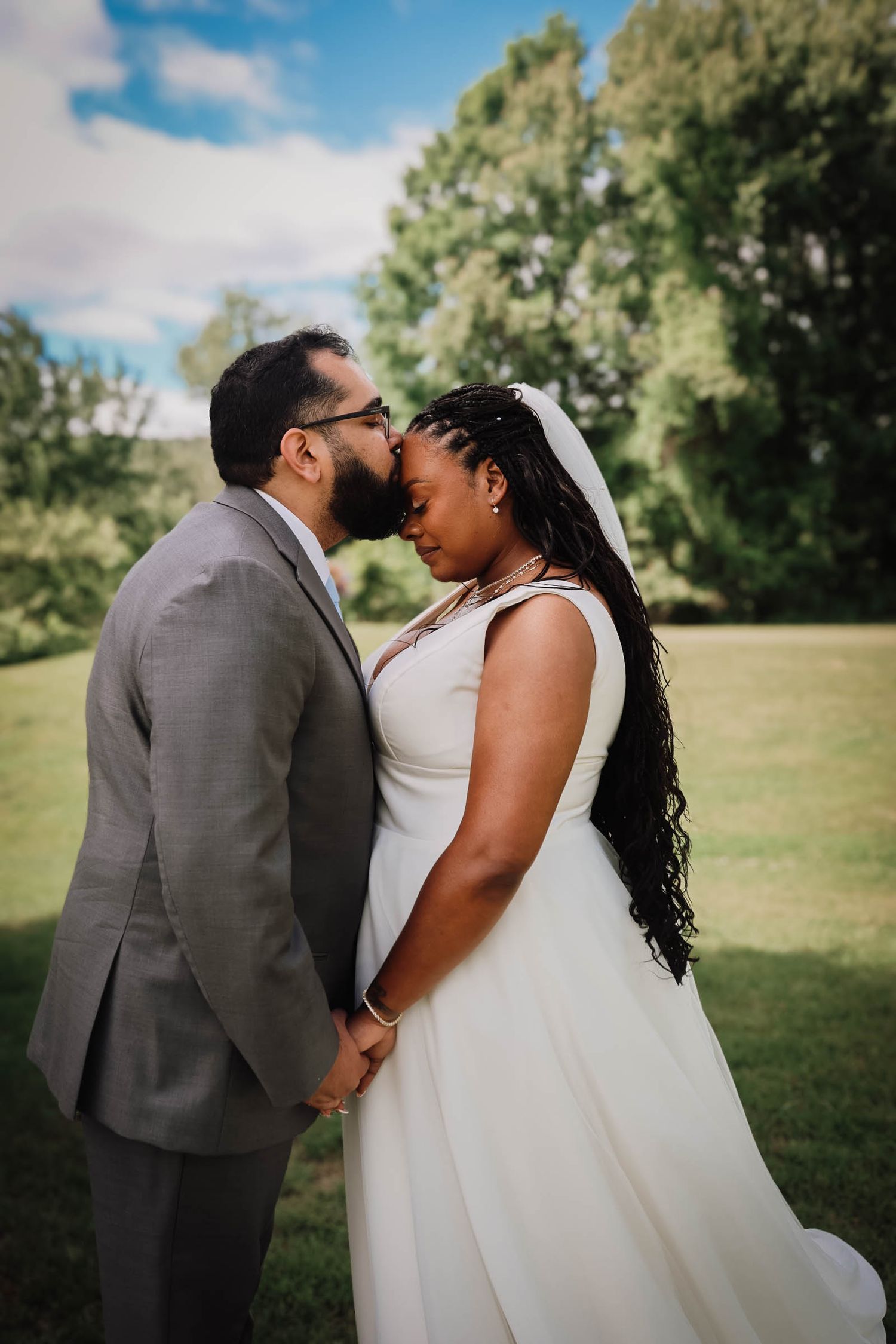 Bride and groom sharing tender forehead kiss outdoors on lush green lawn on their wedding day.
