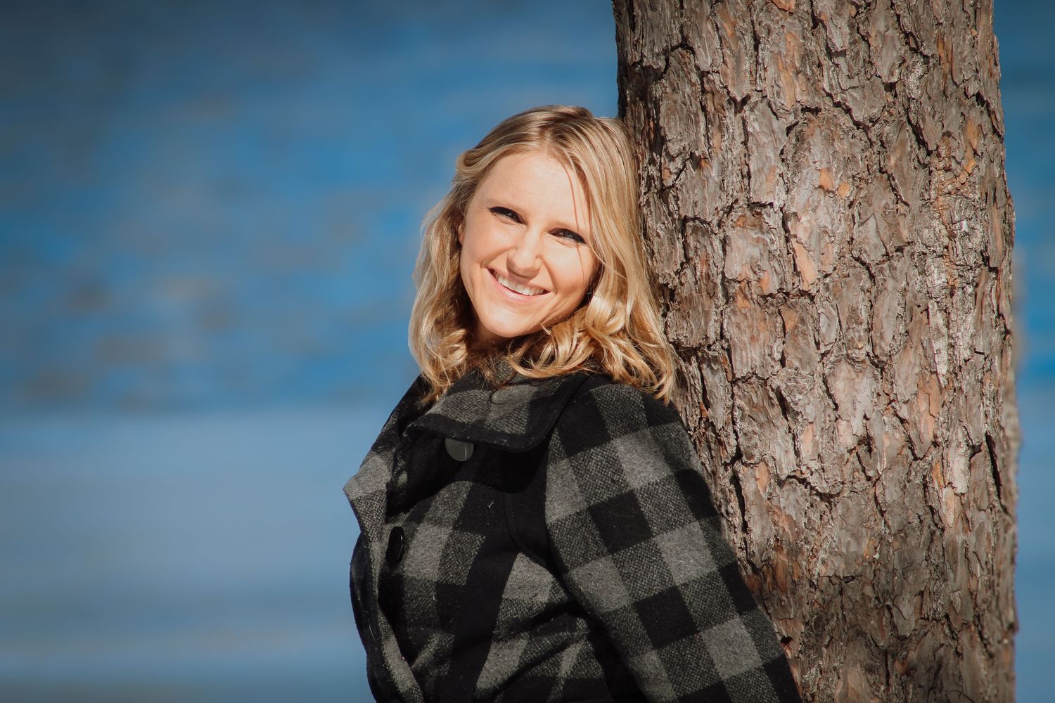 A person wearing a black and gray buffalo plaid jacket smiles while leaning against a large pine tree trunk.