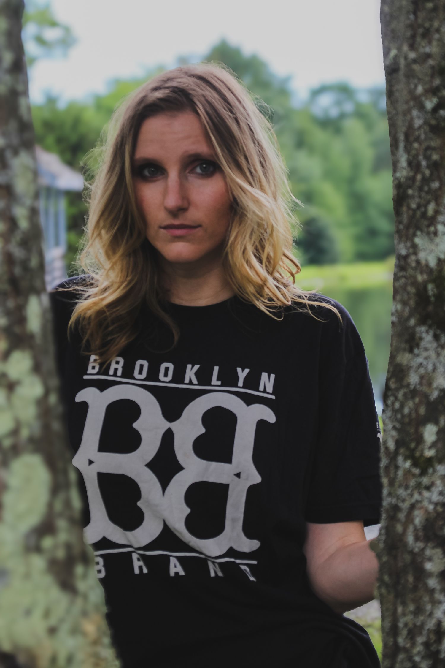 Person wearing black Brooklyn t-shirt poses between trees in outdoor setting.