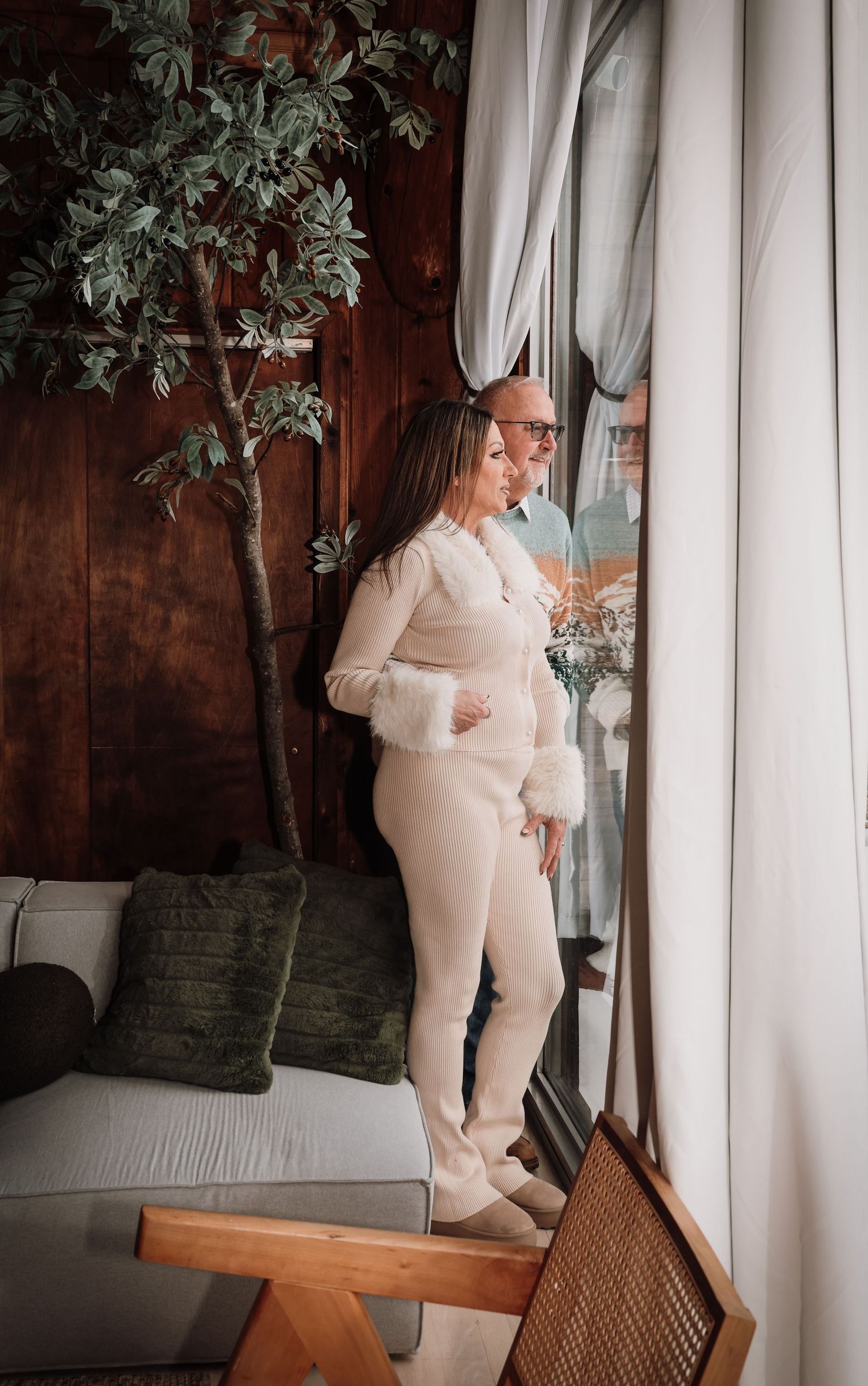 A figure in a cozy cream-colored loungewear set stands near white curtains and indoor plants in a home setting.