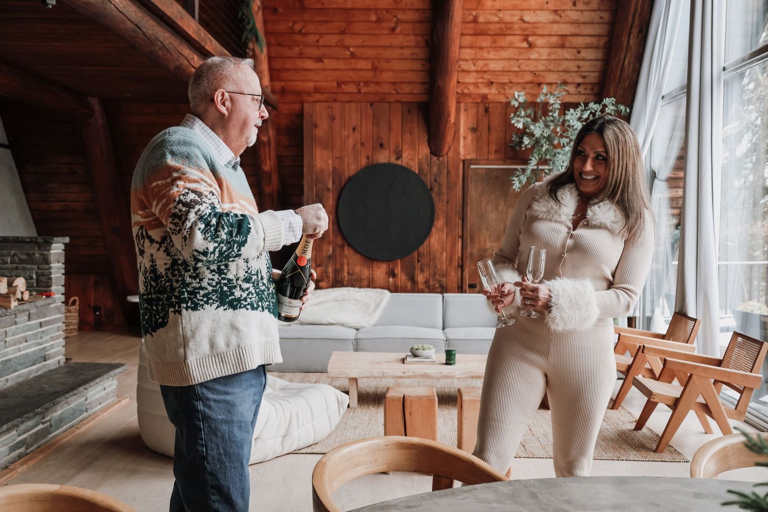 Two people enjoying drinks and conversation in a cozy wooden cabin interior with natural lighting by large windows.