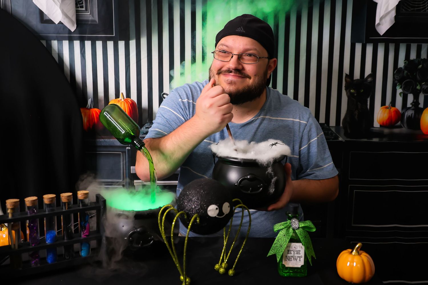 A person sits at a Halloween-themed table with decorative pumpkins and colorful bottles in a dark room with green lighting.