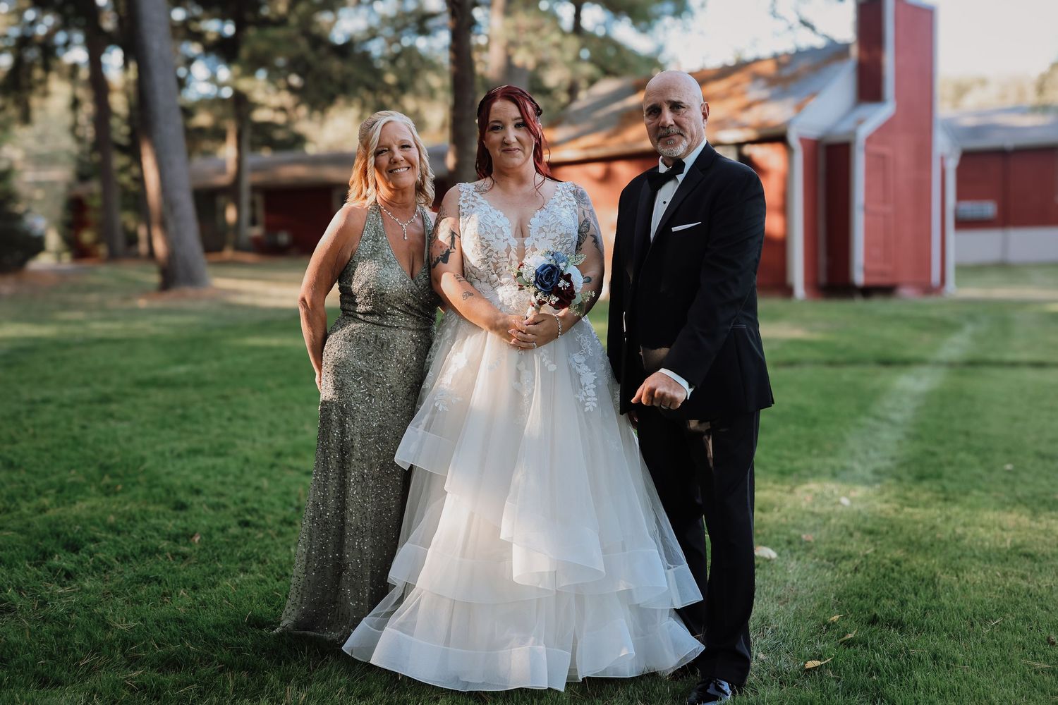 A bride in a layered white wedding gown poses with two guests on a lawn with a red barn in the background.