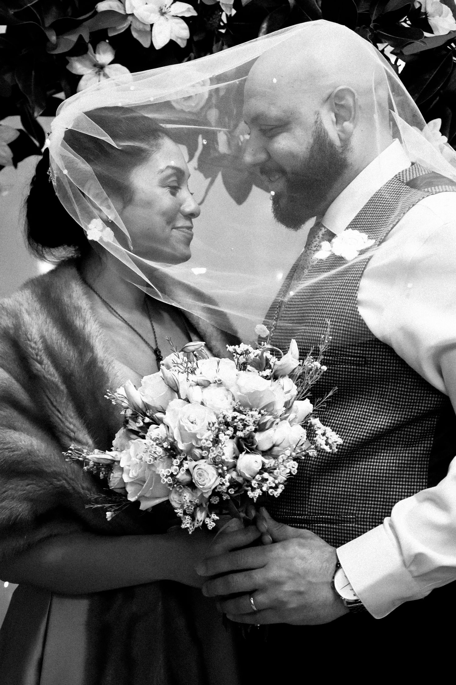 A tender black and white wedding moment with a couple sharing an intimate moment under a veil with a white floral bouquet.