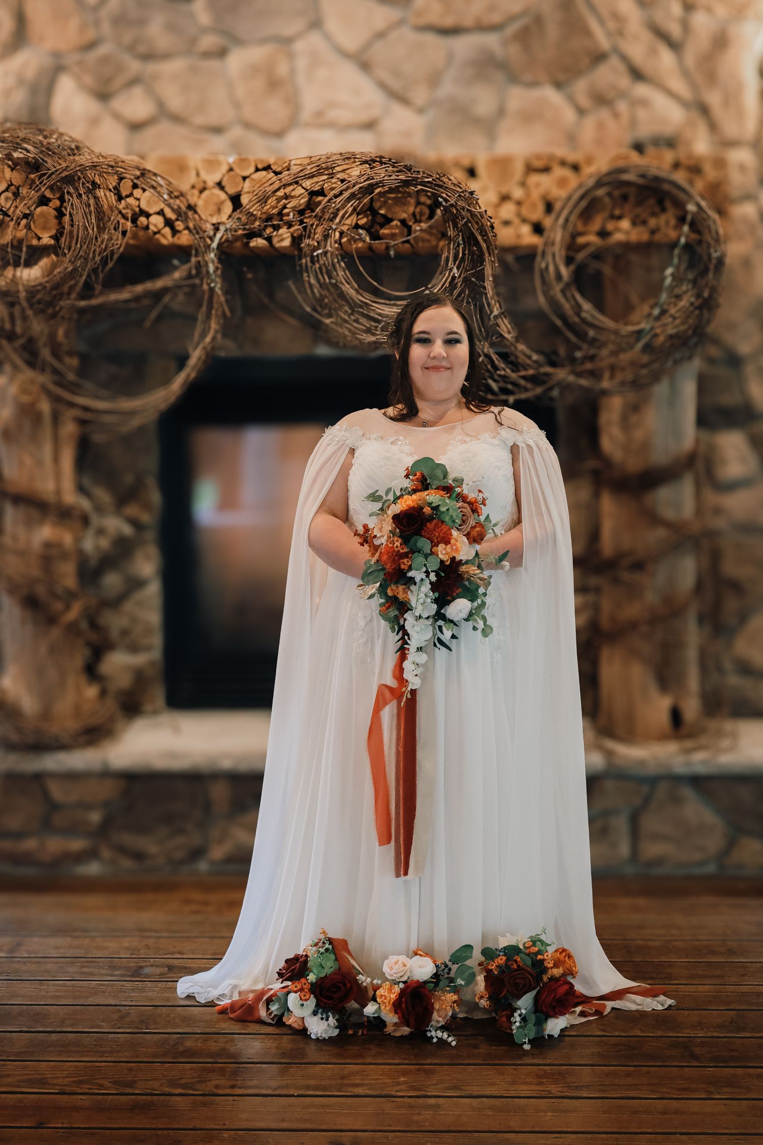 A bride in a flowing white dress holds a vibrant red and white bouquet against a rustic stone wall backdrop.
