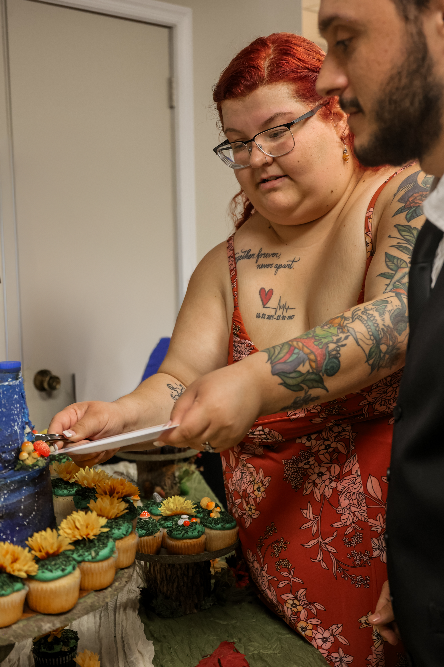 A person with tattoos in a red floral dress arranges yellow flowers and greenery on a table.