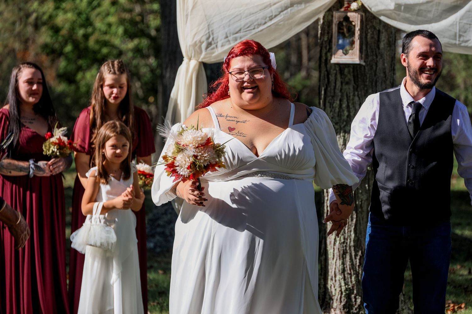 Wedding party stands together outdoors under white fabric draping with burgundy and white attire and autumn bouquets.